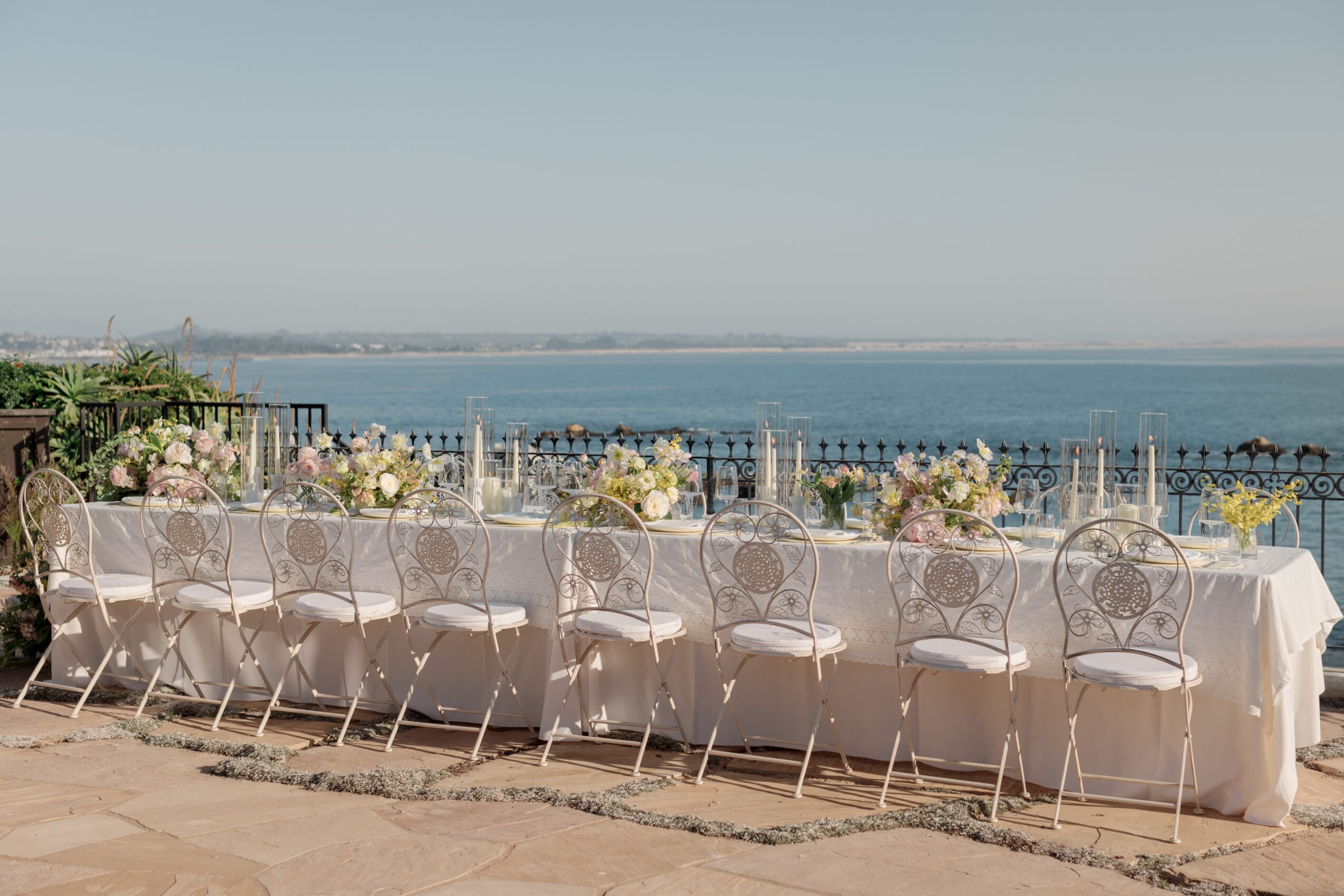 Long table with chairs outdoors overlooking water, decorated with flowers and tableware, under a clear sky.