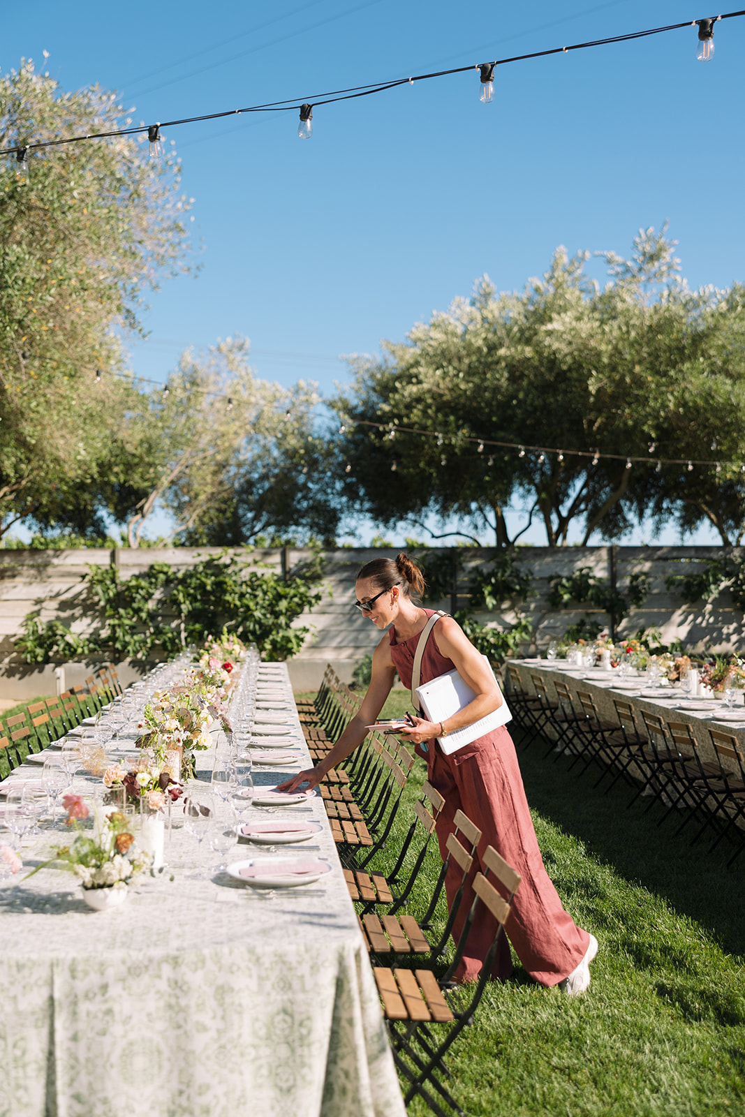 Woman in a white top and long skirt setting a table outdoors with flowers and tableware, string lights above, trees in background.