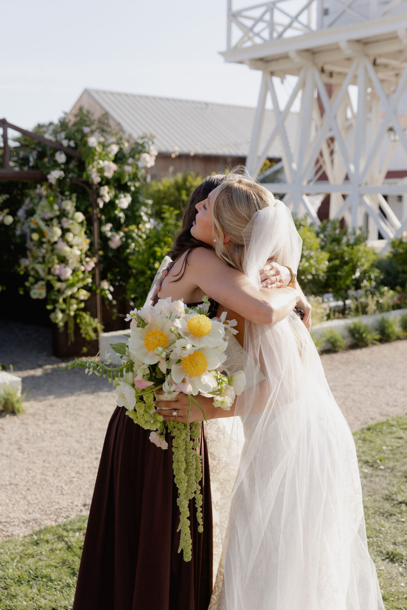 Two women in wedding dresses embrace outdoors, holding a bouquet of white and yellow flowers, with a garden and white structure in background.
