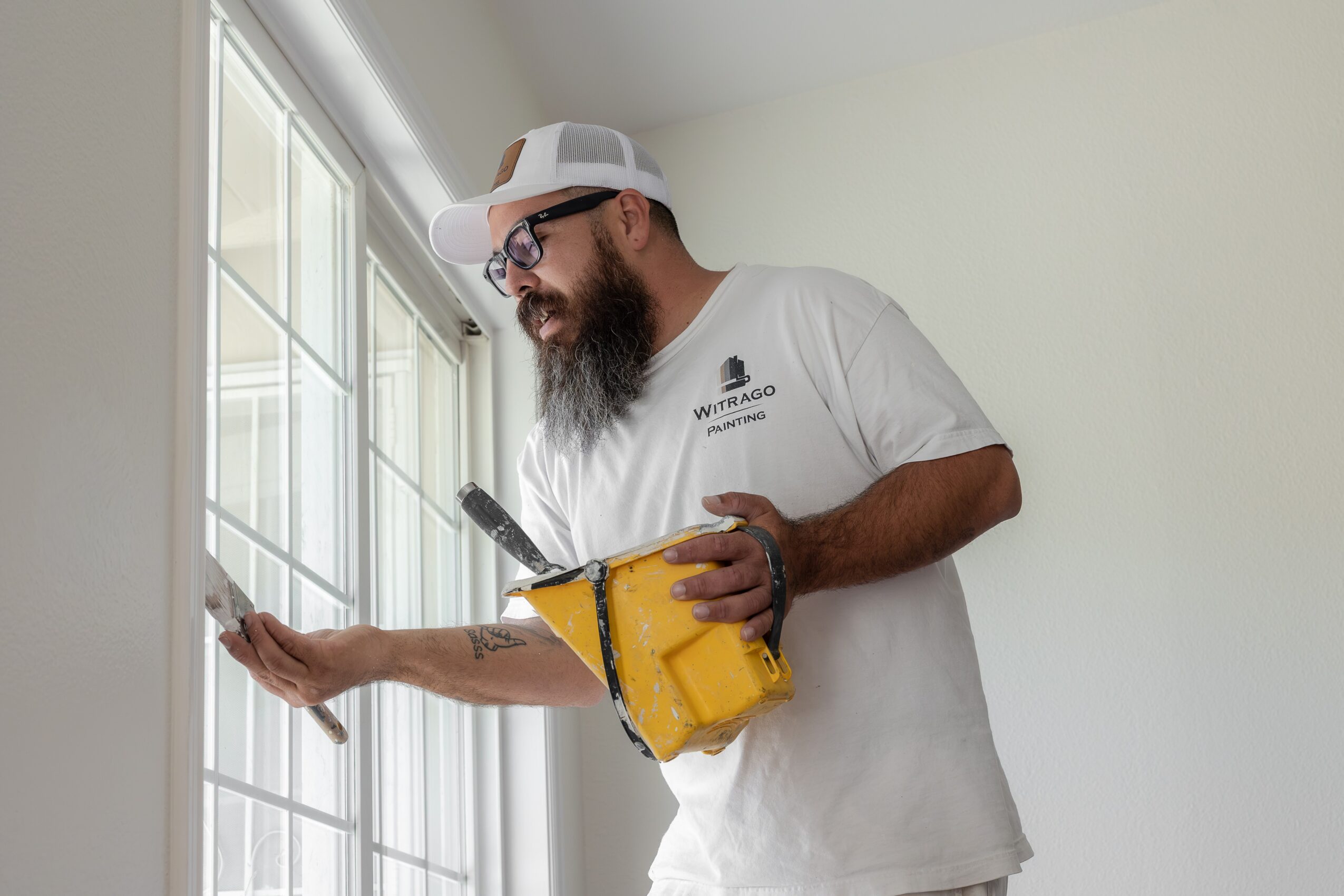 Man with a beard and glasses cleaning a window with a squeegee, holding a yellow bucket, wearing a white cap and shirt.