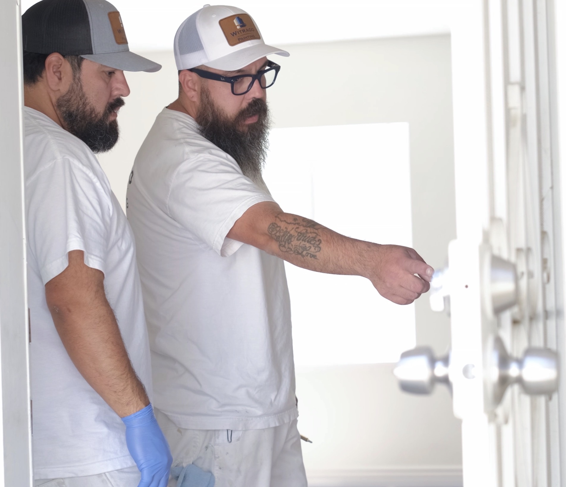 Two men in casual clothing and hats look at a whiteboard or display, one pointing at it, in a bright room.
