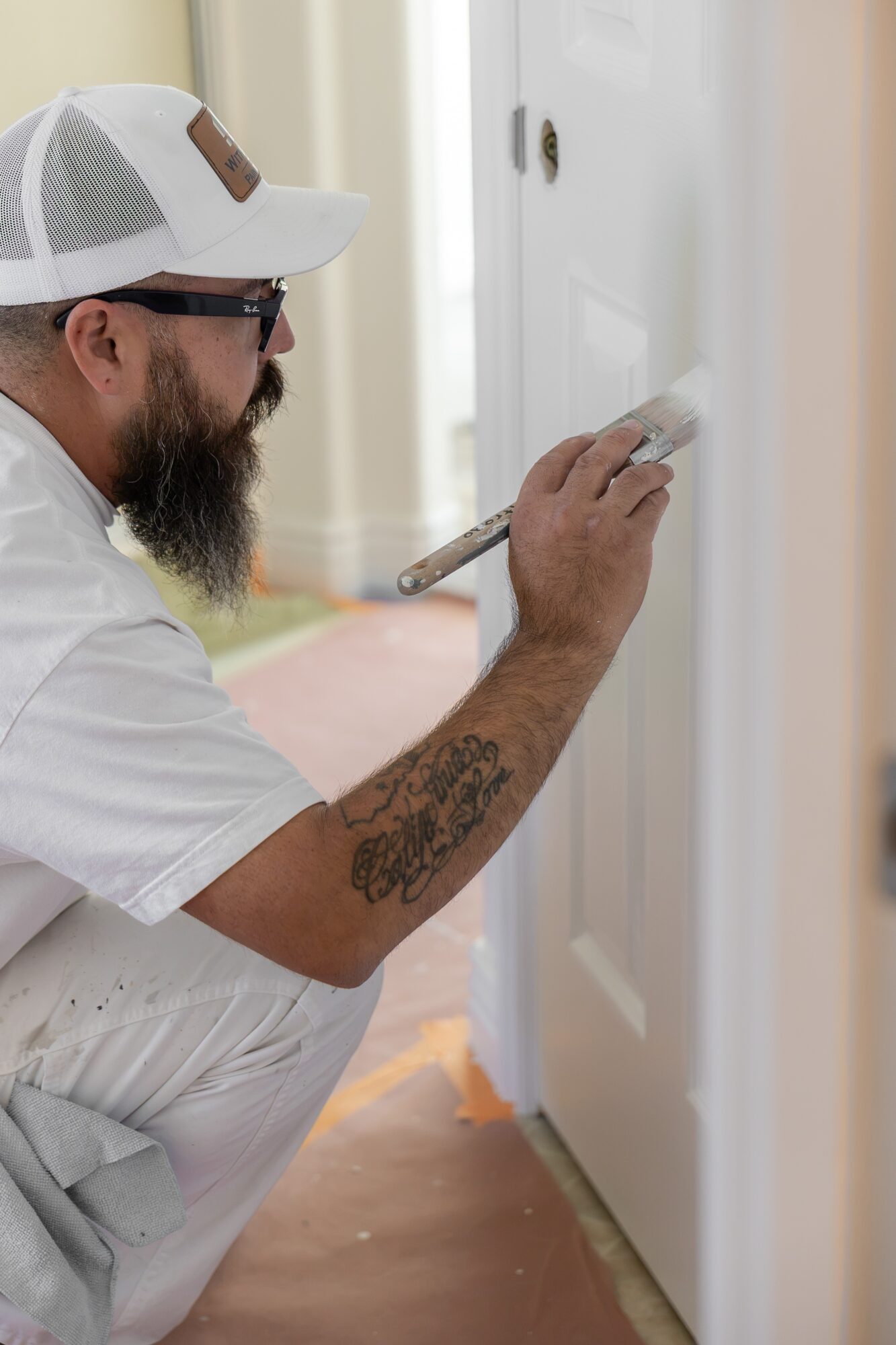 Man with a beard, wearing a white cap and glasses, painting a door with a brush, kneeling on the floor.
