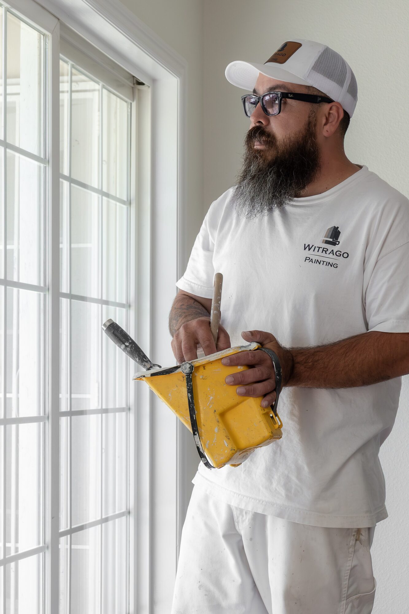 Man with a beard and glasses wearing a white cap and T-shirt, holding a paint roller and a yellow paint tray, standing near a window.