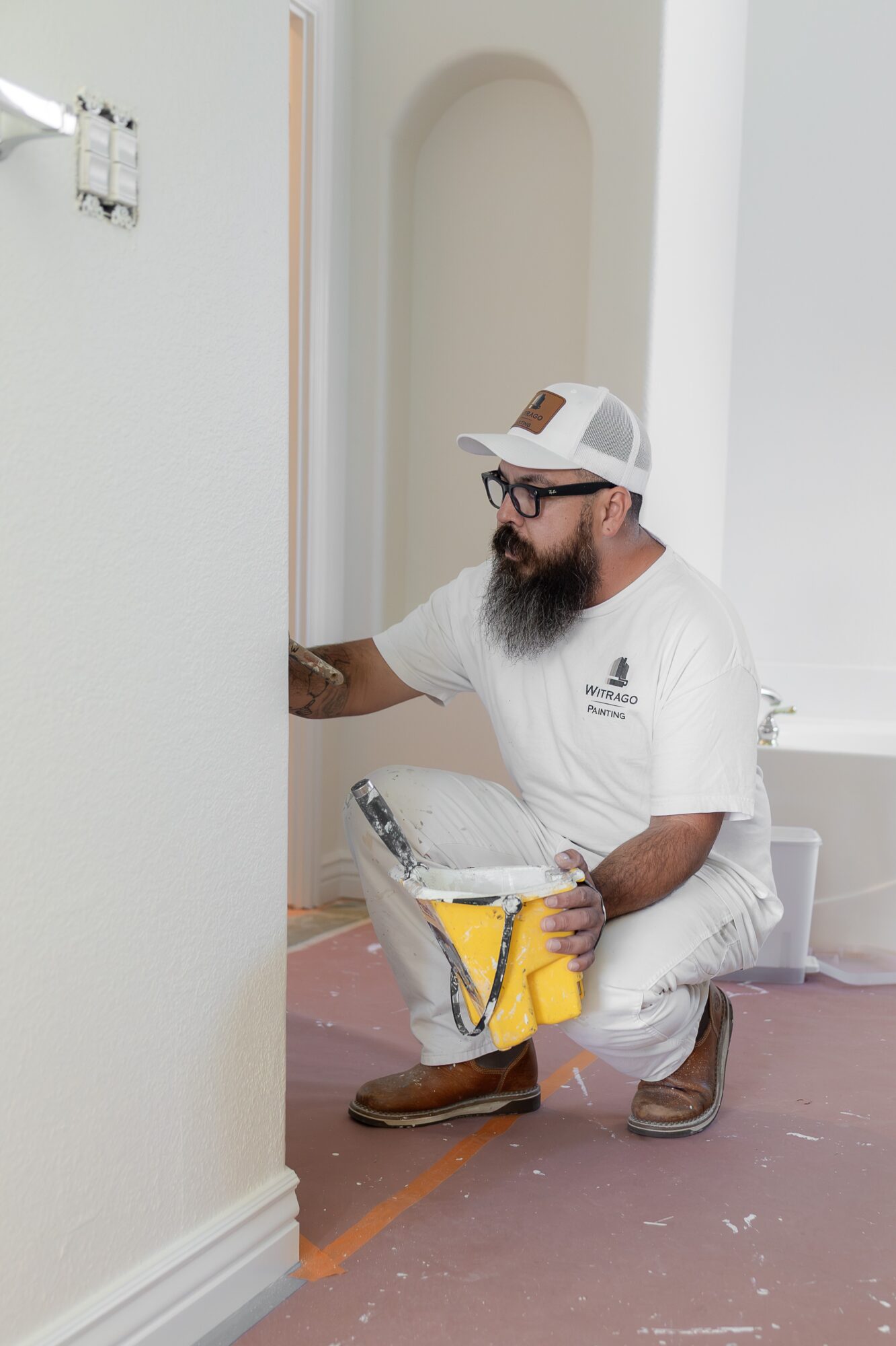 Man kneeling on the floor, working on a wall with tools, wearing a white cap, glasses, and white clothing.
