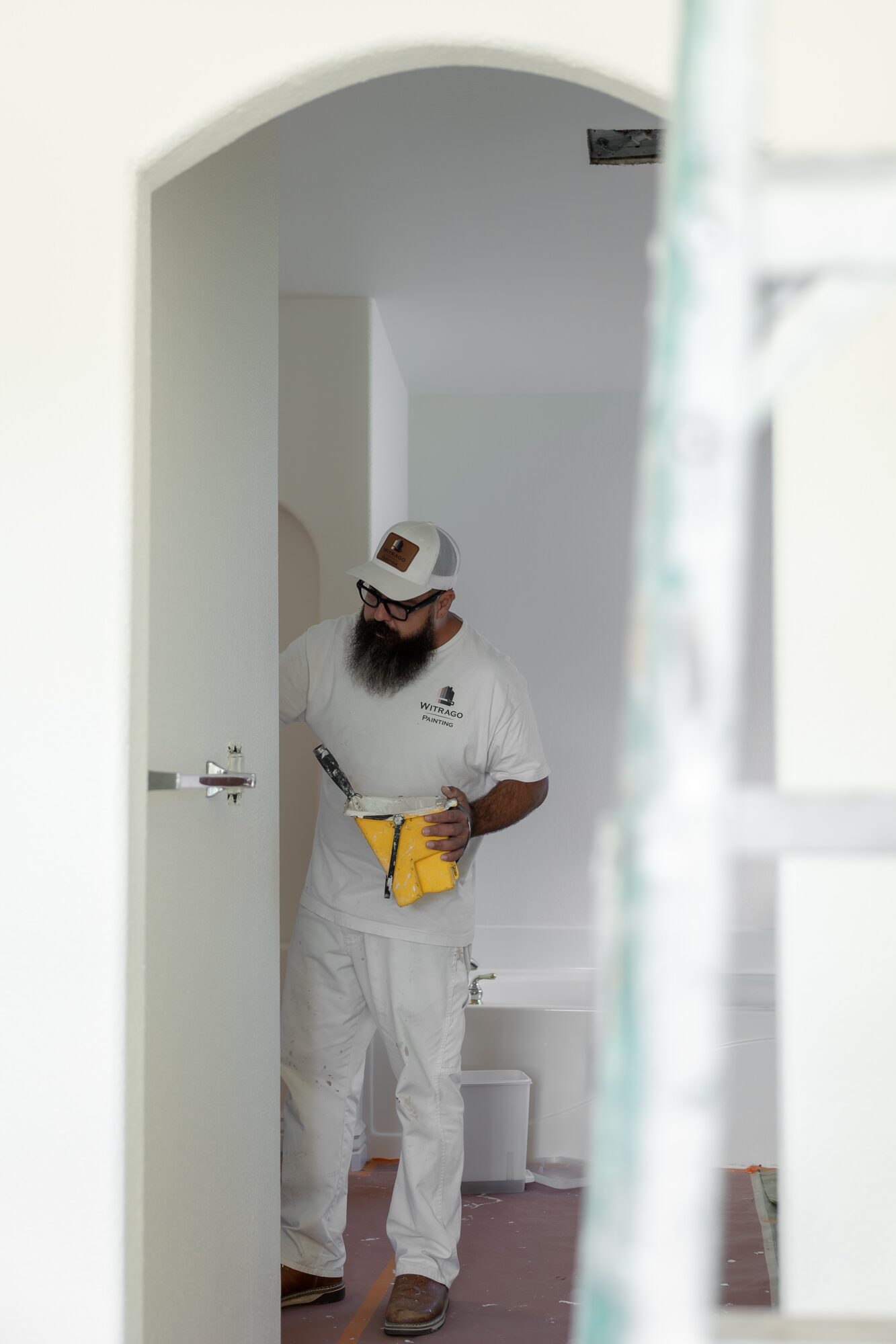 Person with a beard wearing white clothing and a cap, standing in a doorway, holding a yellow object.