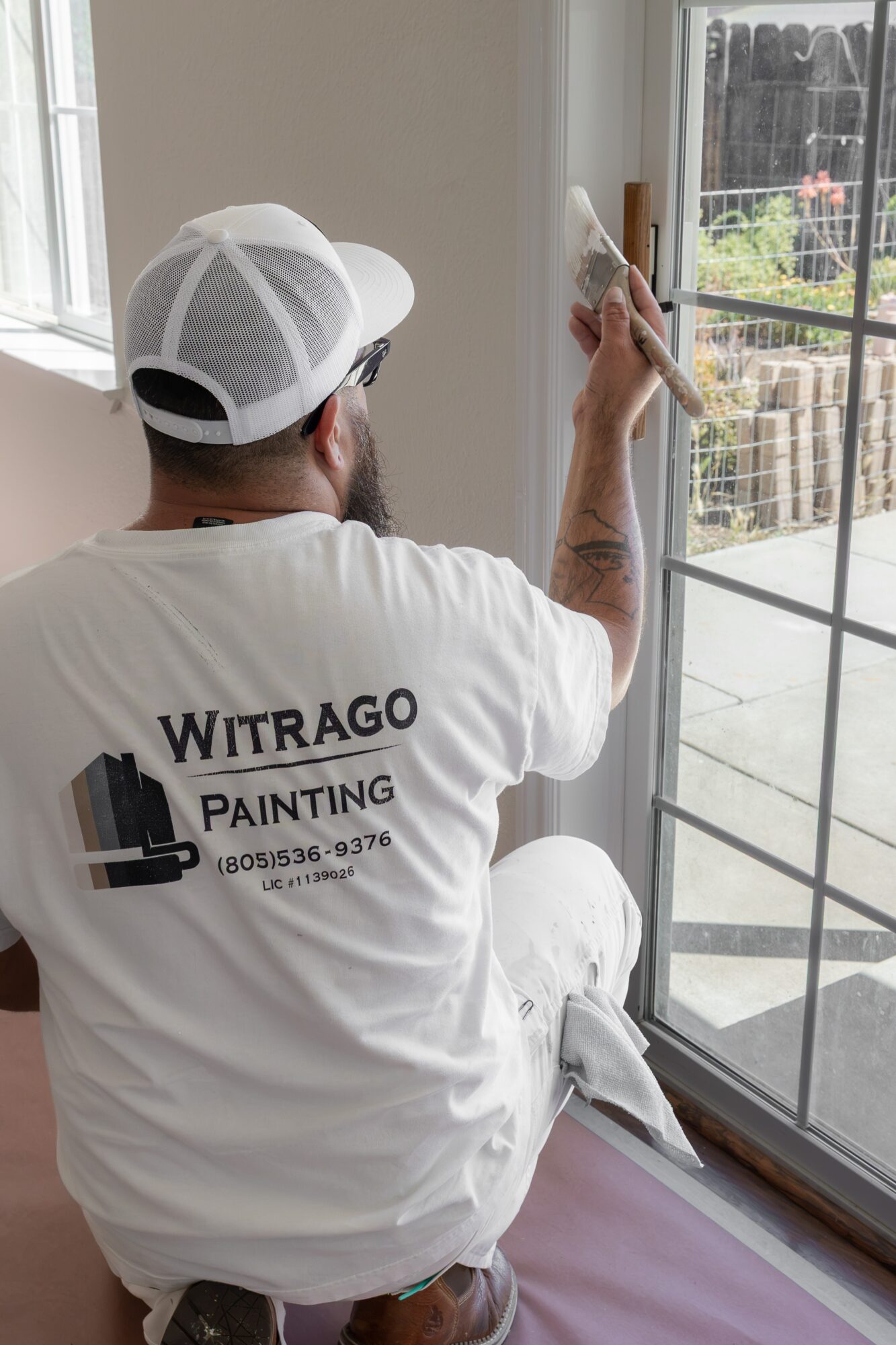 Person painting window frame inside a room, wearing a white cap and shirt with company logo, sitting on a stool.