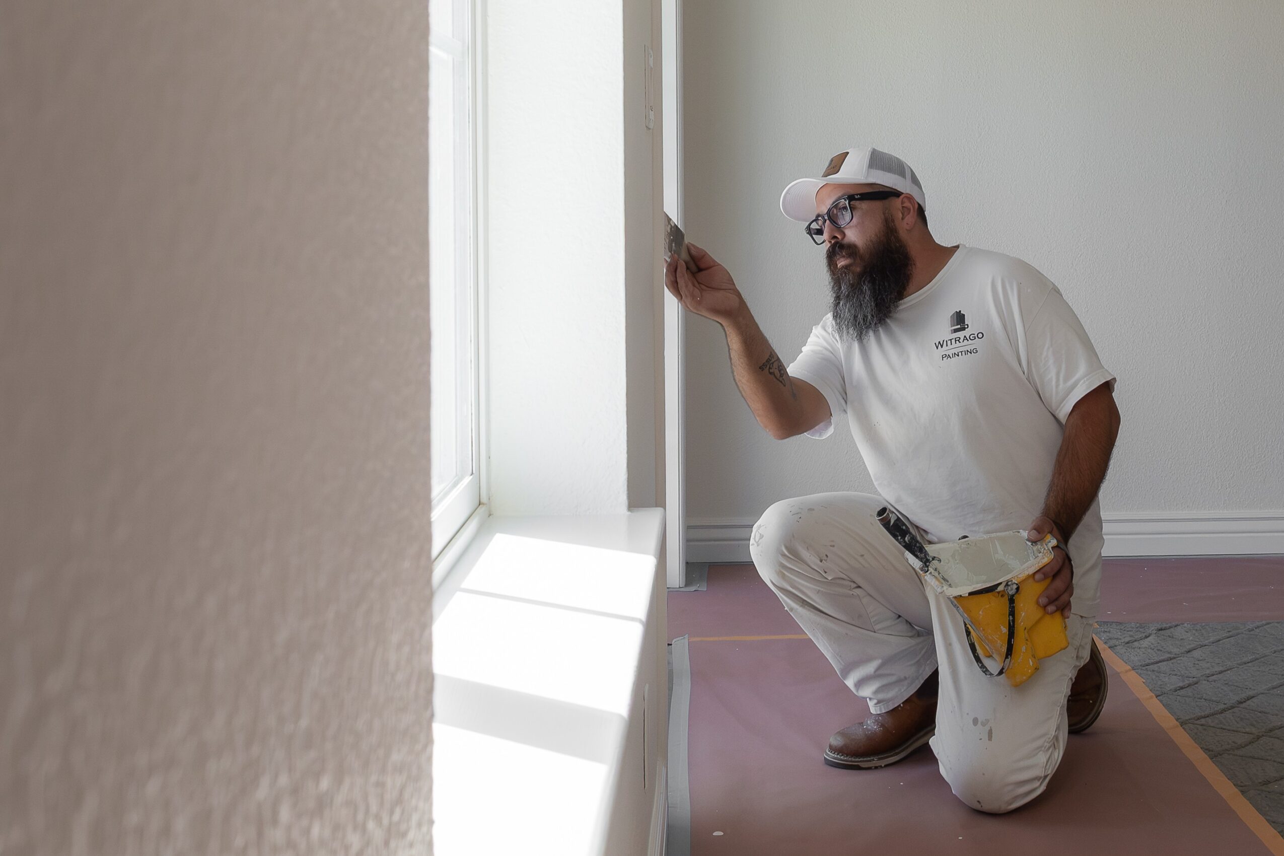 Man with a beard wearing a white cap and t-shirt kneeling near a window, looking and pointing at the window, with sunlight coming in.
