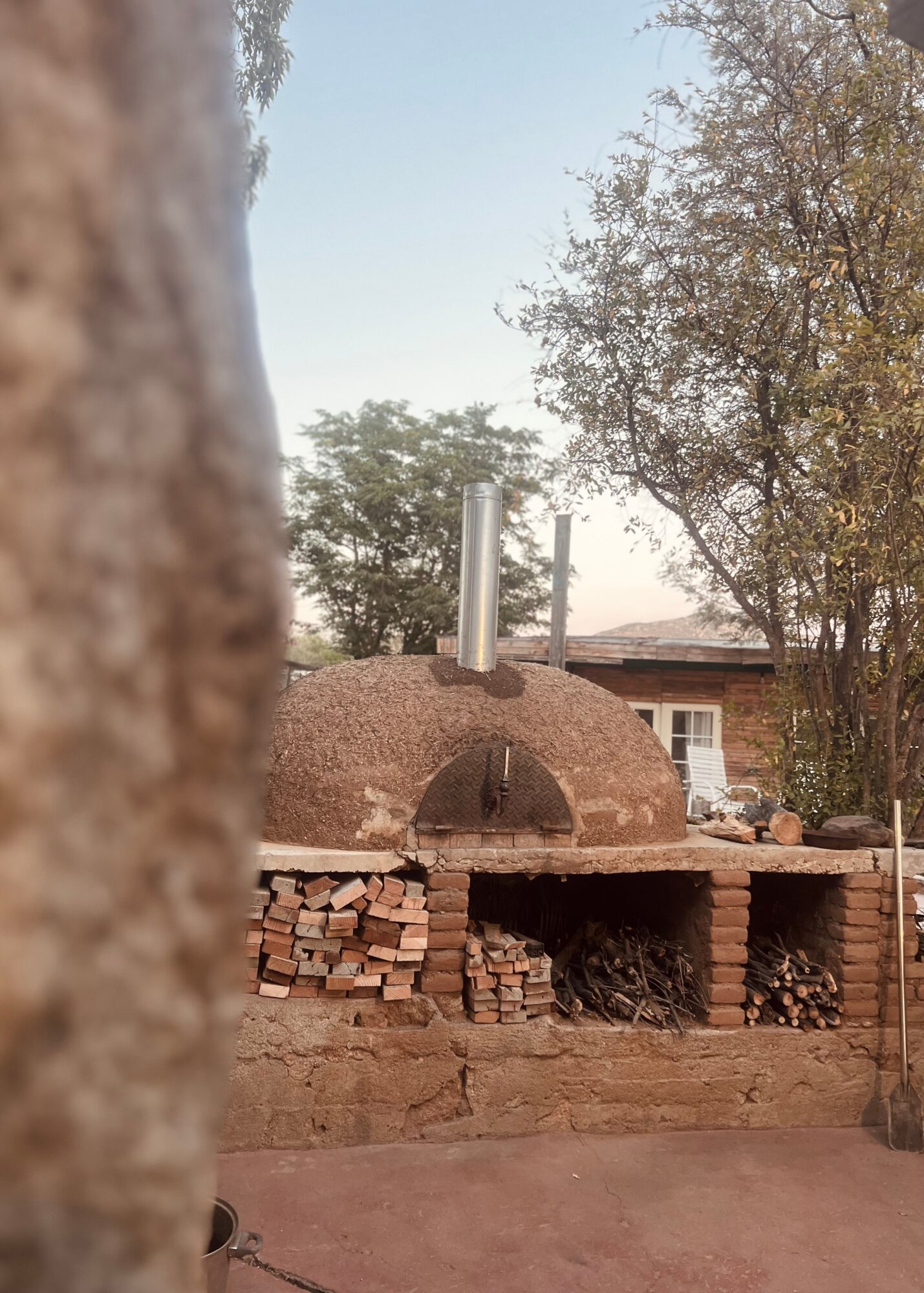 Outdoor brick oven with chimney pipes, surrounded by trees and a house in the background.