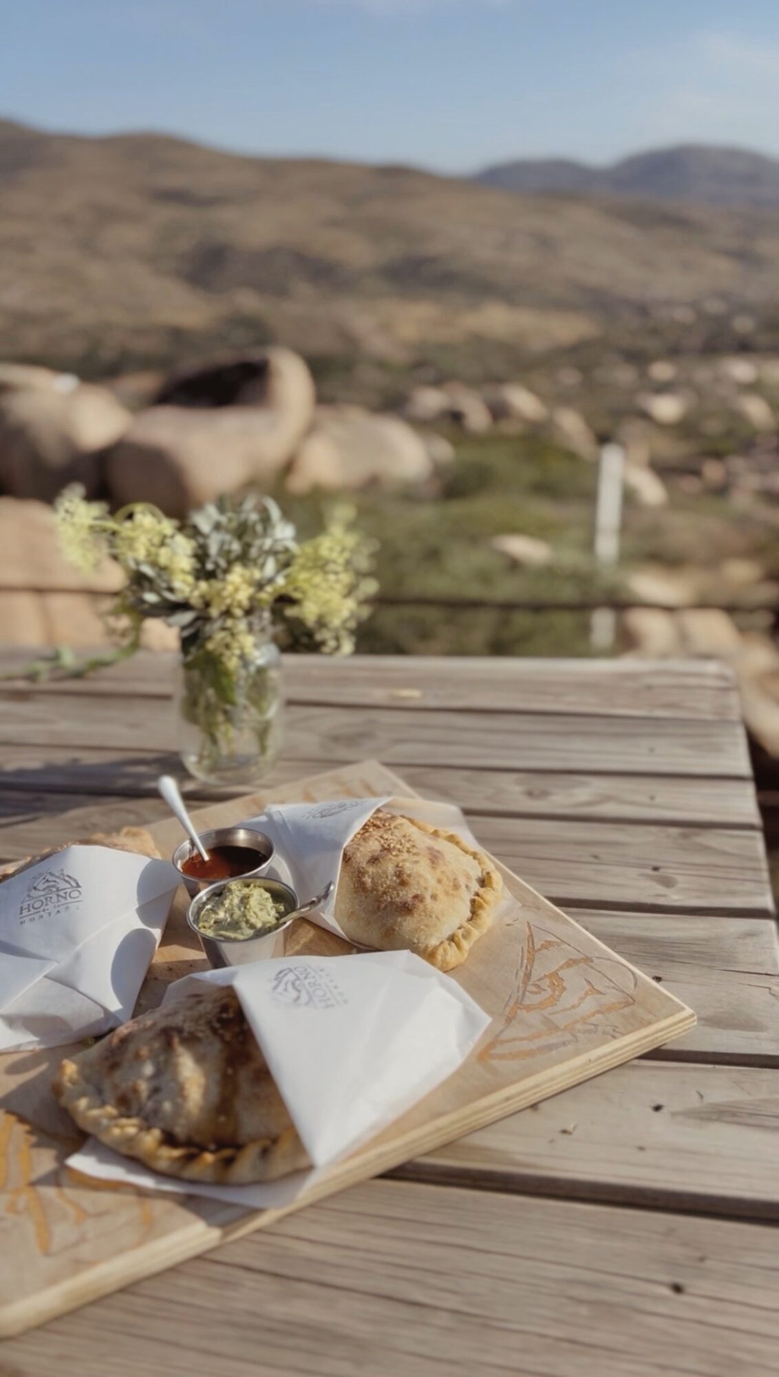 Wooden table with baked goods, a jar of flowers, and a small bowl, outdoors with hills in the background.