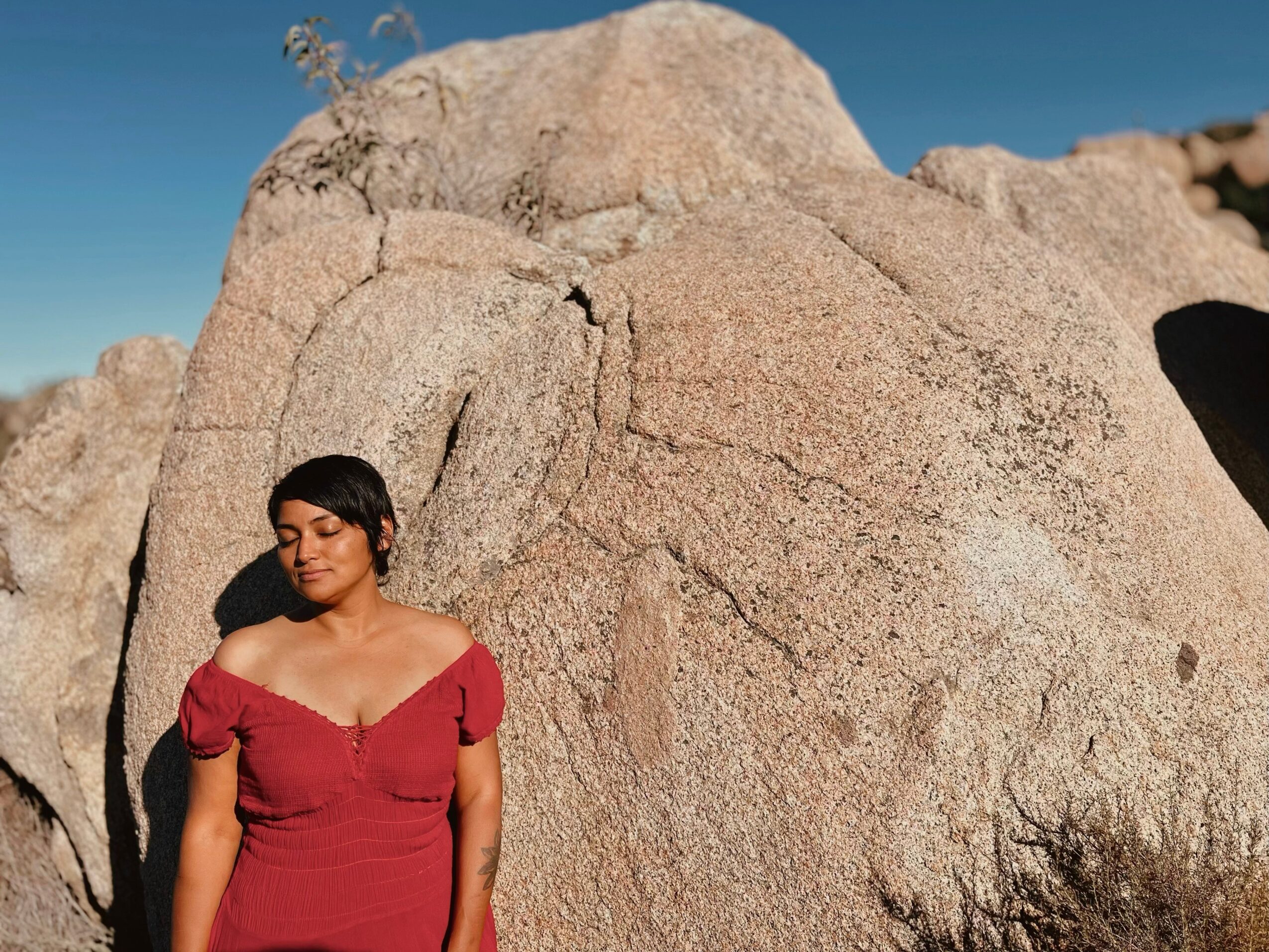 Woman in red dress standing next to large rock formation outdoors under clear blue sky.