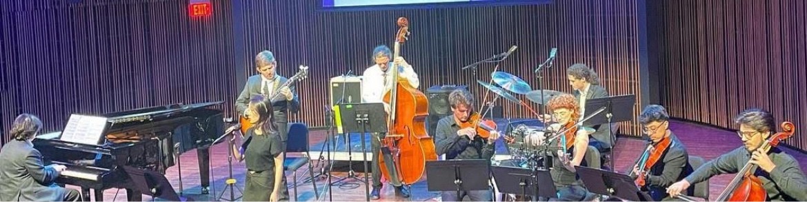 Group of musicians performing on stage with various instruments, including piano, guitar, and drums, in front of a purple curtain.