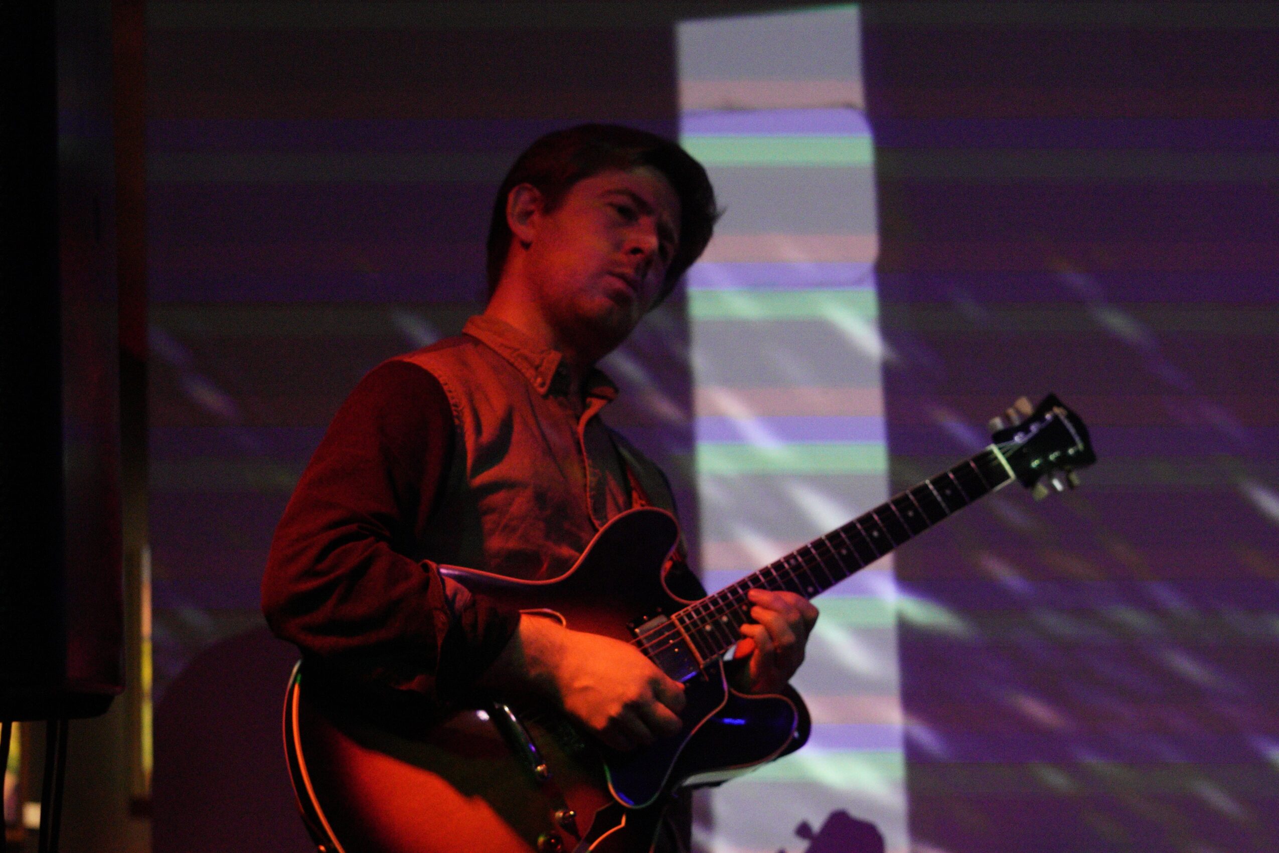 Person playing guitar on stage with colorful background, wearing a dark jacket, looking to the side.