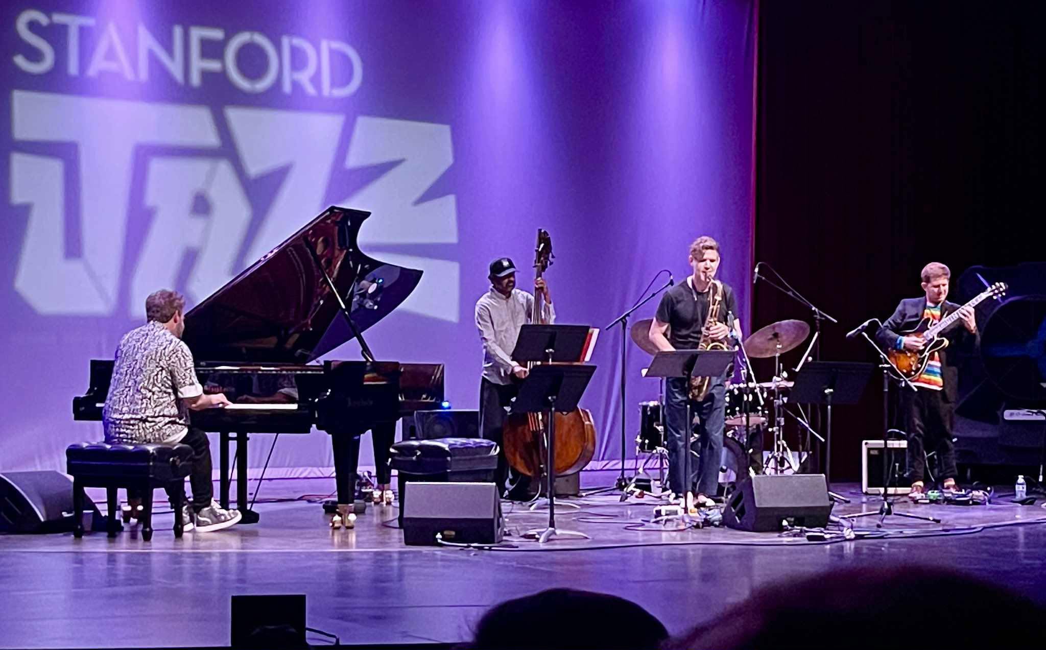 Jazz band performing on stage with a pianist, double bassist, saxophonist, and guitarist, under purple lighting, with 'STANFORD' visible in the background.