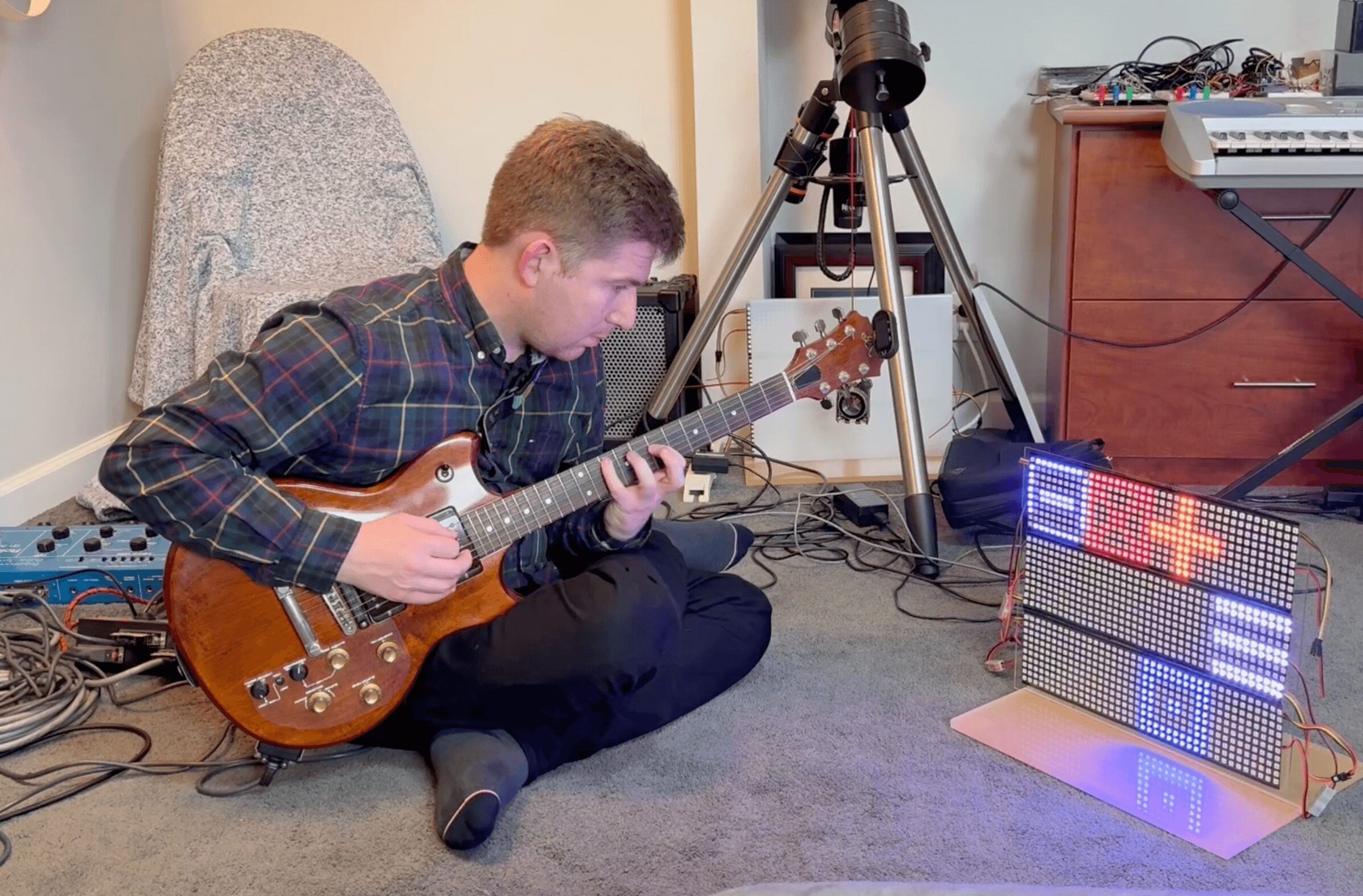 Person sitting on floor playing electric guitar, surrounded by camera equipment and electronic devices, in a room.