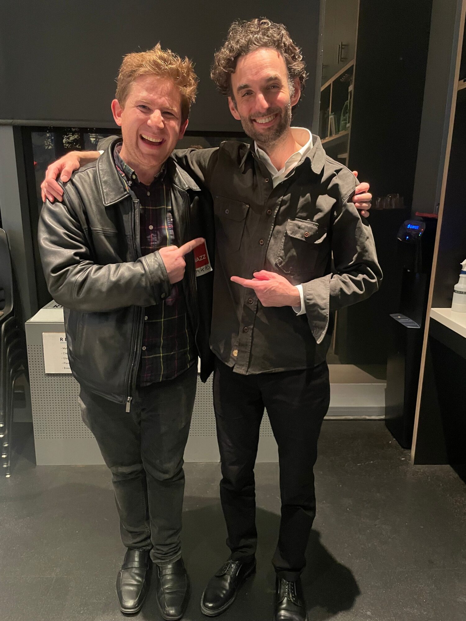 Two men standing indoors, one pointing at the other, both smiling, with dark flooring and a black wall behind them.