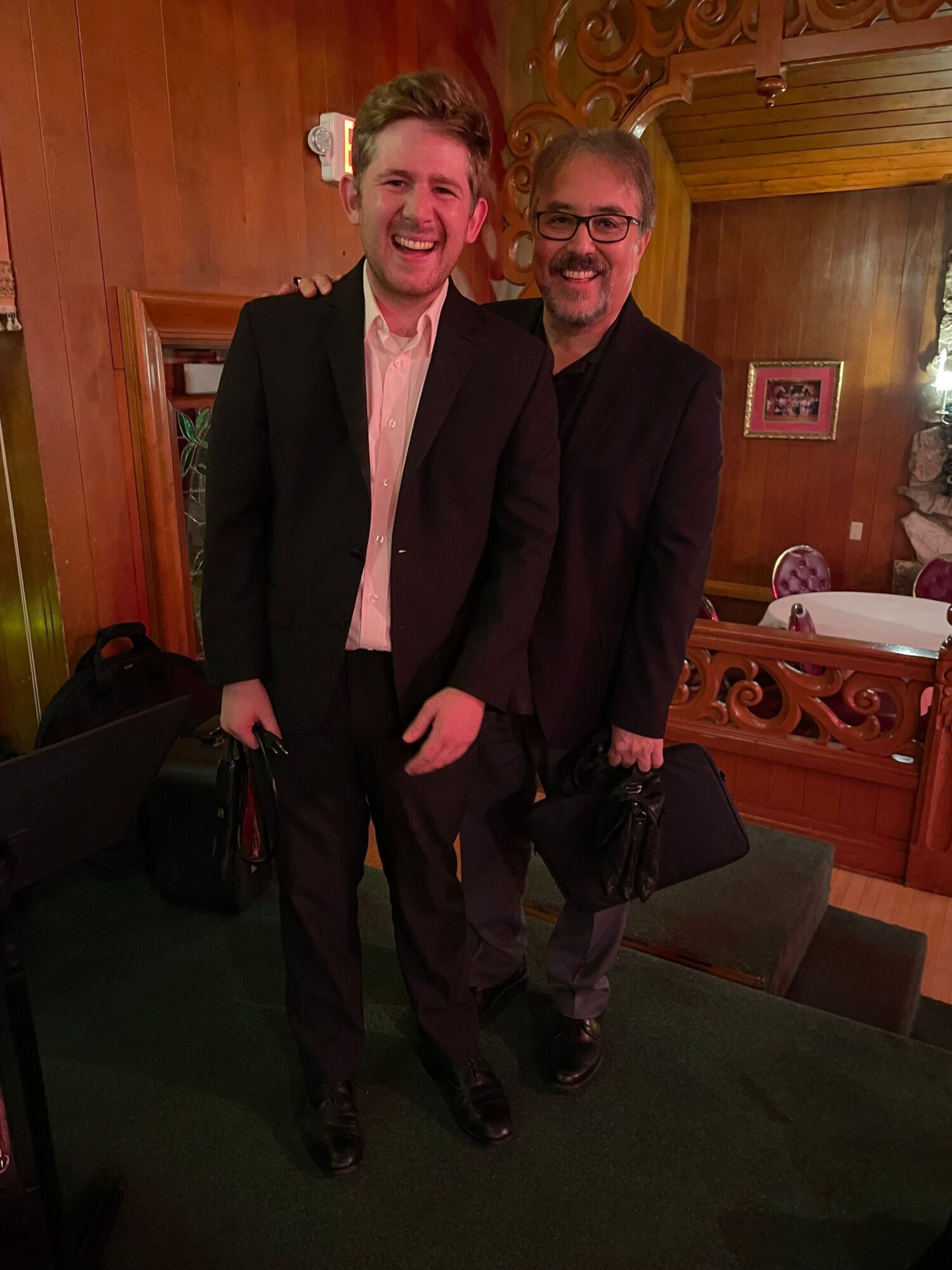 Two people in formal attire standing indoors, one with glasses and the other with a pink shirt, smiling.