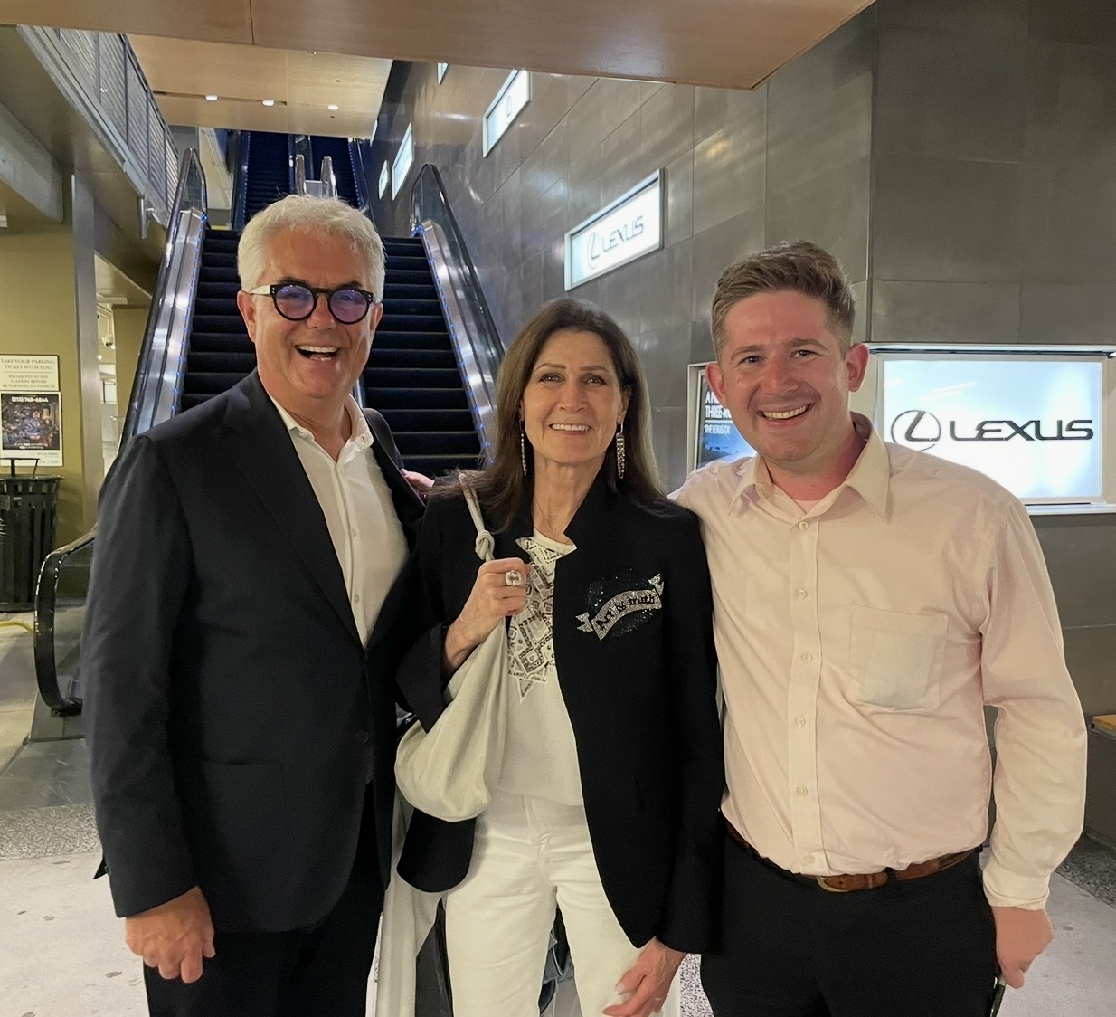 Three people smiling indoors, two men and one woman, standing in front of escalators and Lexus signs.