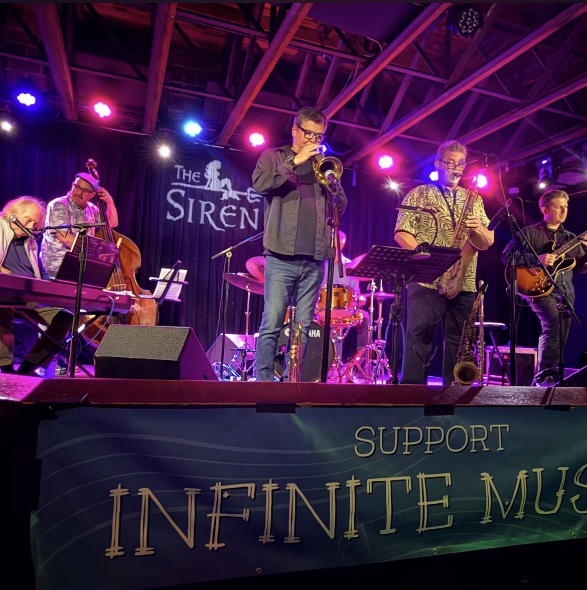Band performs on stage with colorful lighting, supporting Infinite Music, with five musicians playing instruments and singing.