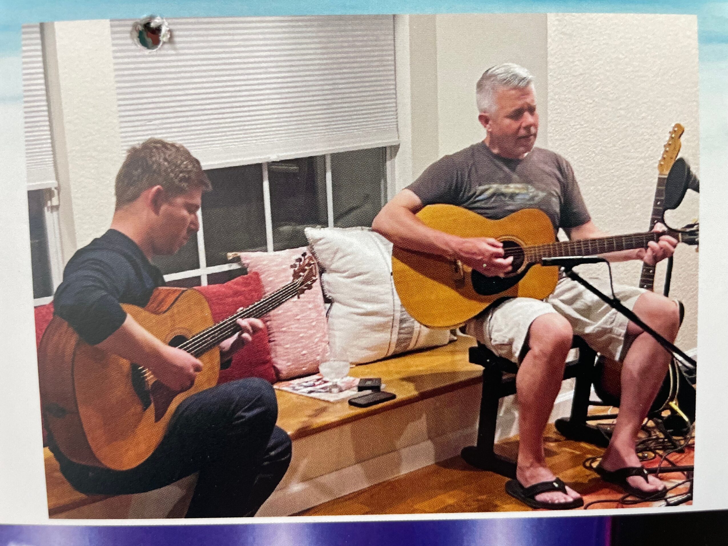 Two people playing guitars on a window seat with pillows, one young and one older, in a room with a window and blinds.