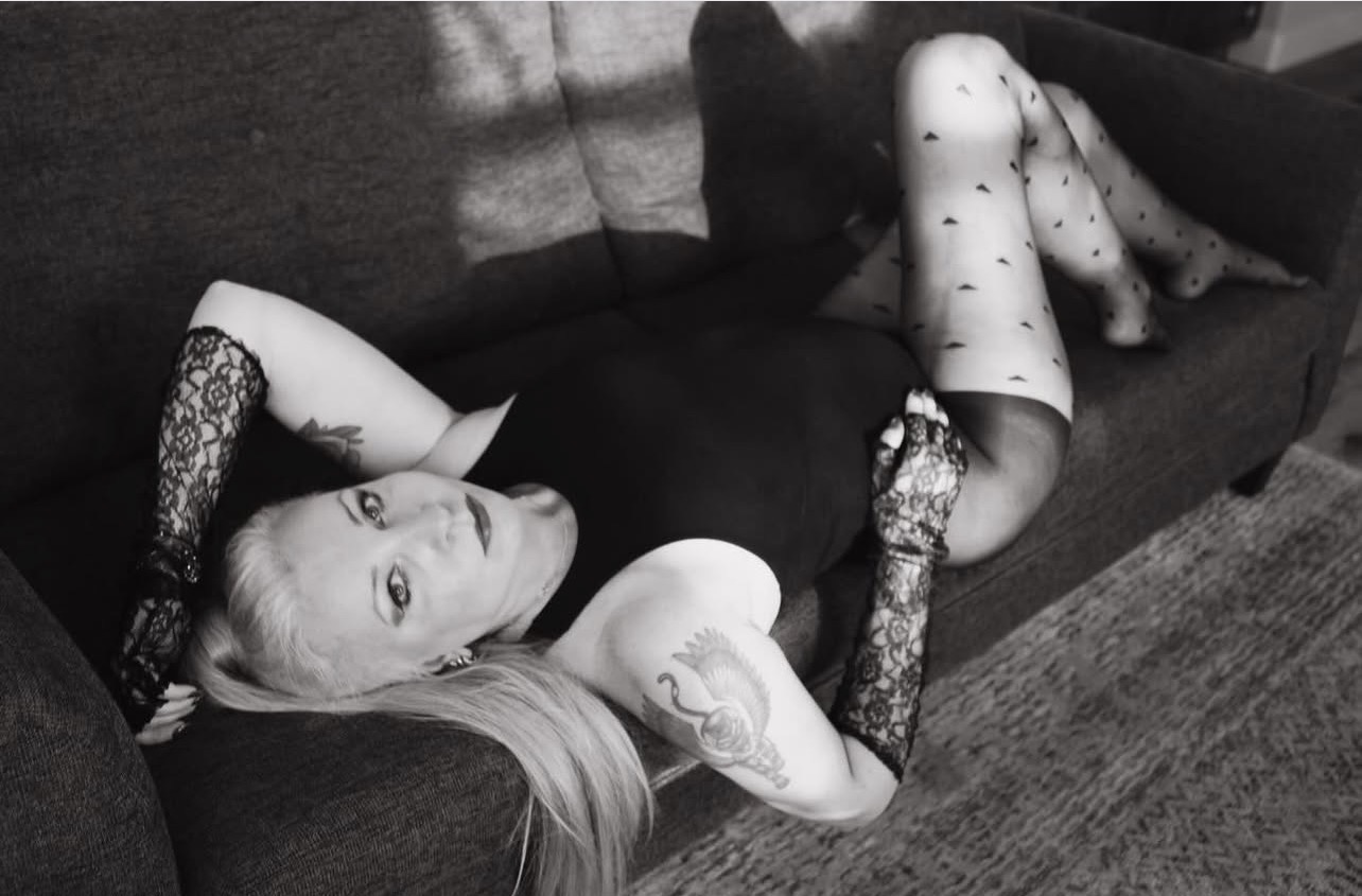 Woman lying on a sofa wearing a black dress and patterned stockings, looking at the camera.