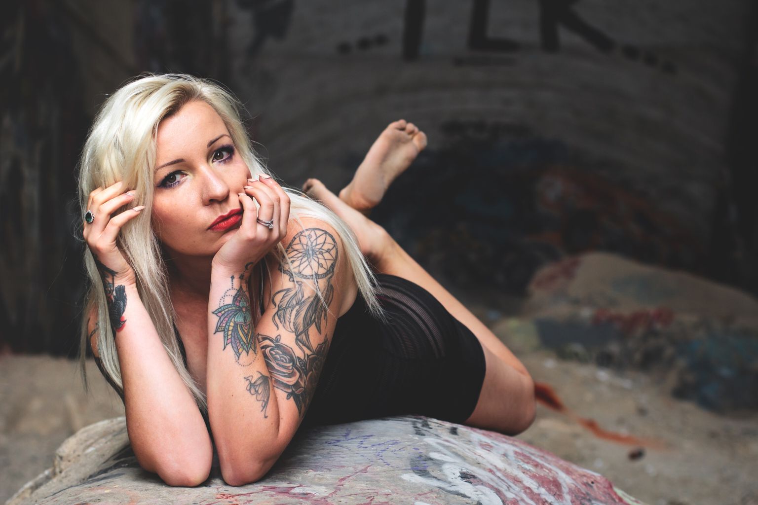 Woman with blonde hair lying on her side, resting her head on her hand, with tattoos on her arm, in a dark setting.