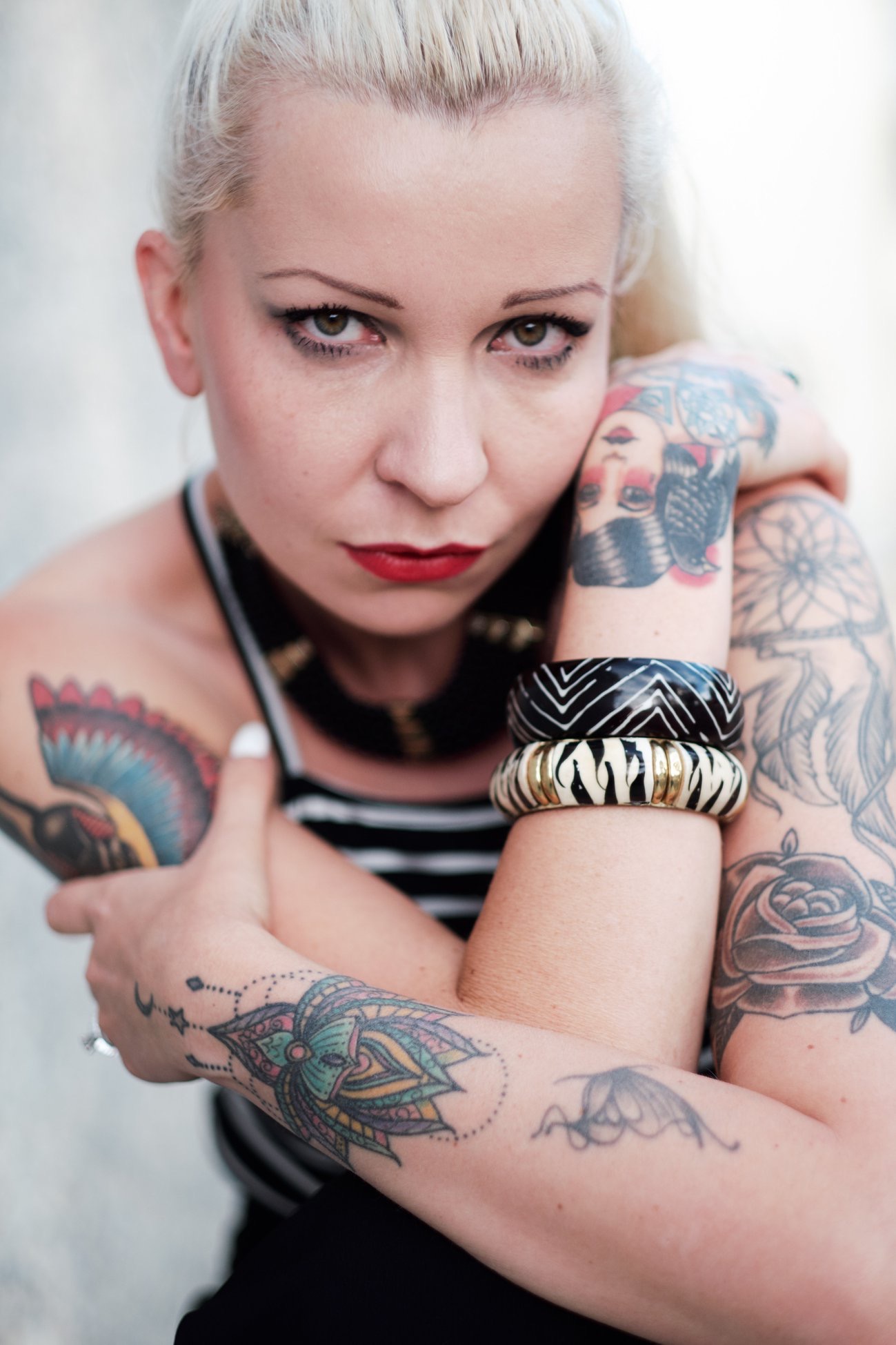 Woman with platinum blonde hair and tattoos on arms, wearing striped top, looking at camera with crossed arms.