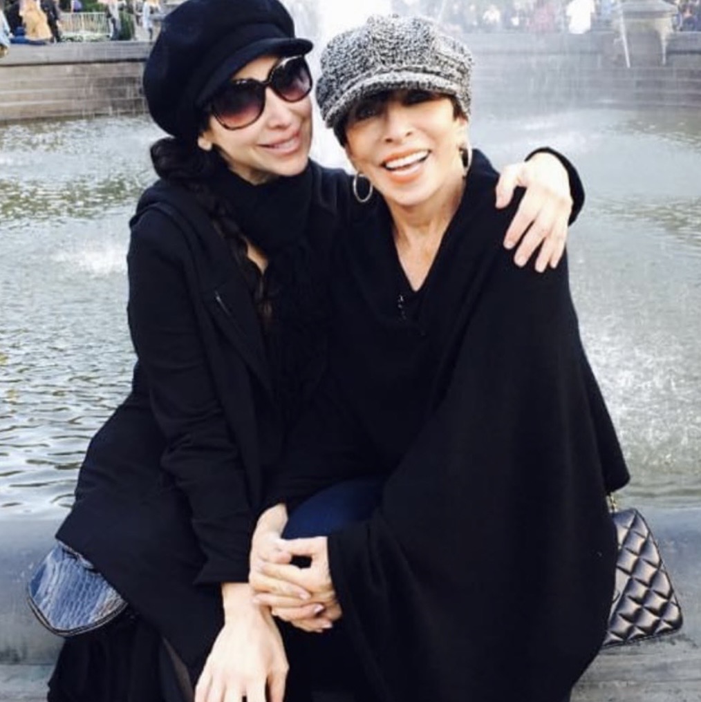 Two women smiling and hugging near a river, wearing hats and dark clothing.