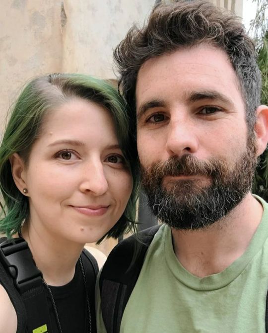 Two people smiling outdoors, close-up portrait, woman with short green hair and man with dark curly hair and beard, both wearing casual clothing.