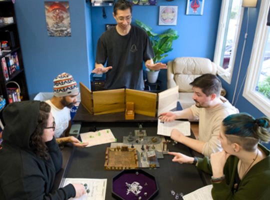 Group of five people playing a tabletop role-playing game around a table with miniatures and books, in a room with blue walls and large windows.