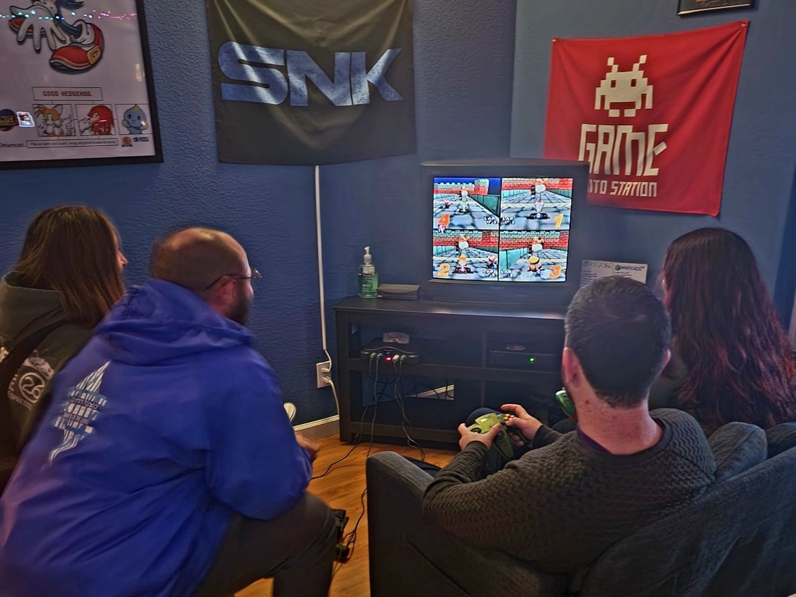 Four people sit in a room watching a video game on a TV, with banners on the wall behind them.