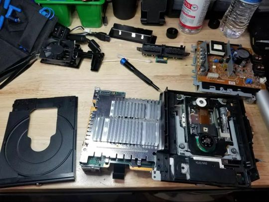 Open computer with internal components, including a cooling system, circuit boards, and a hard drive, on a wooden workbench.
