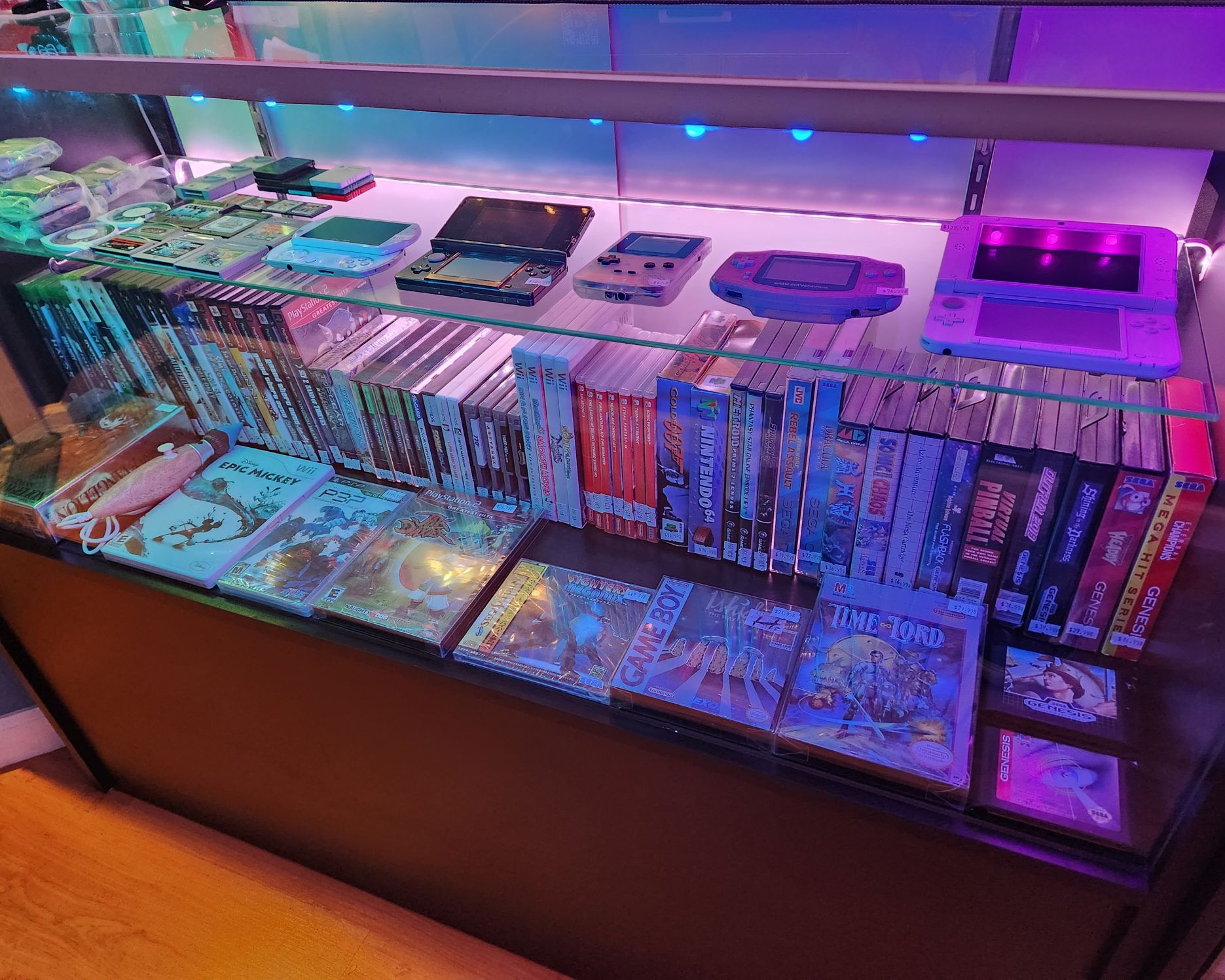 Display case with video game consoles and a collection of video game cases, illuminated by purple and pink lighting.