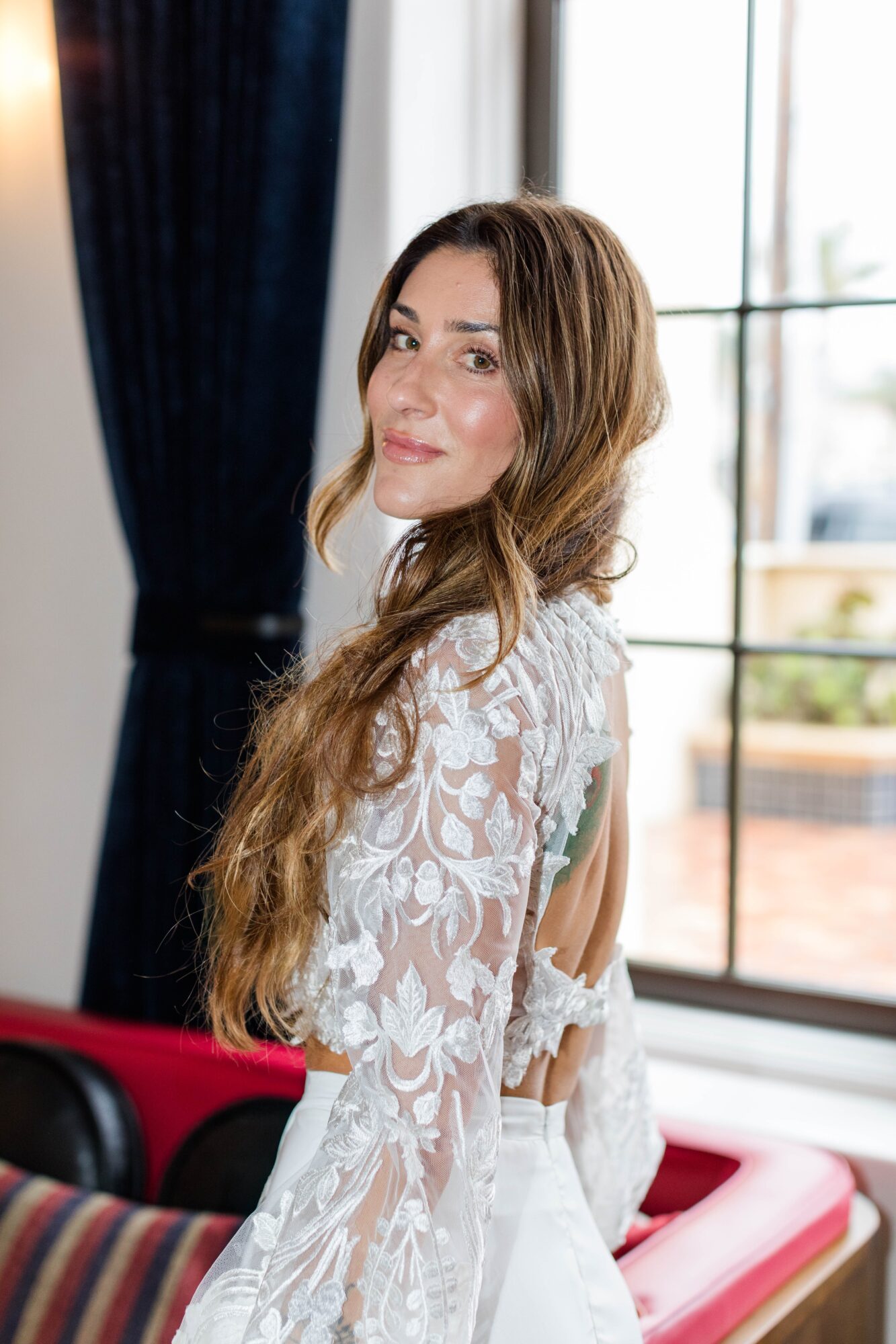 Woman with long hair in a white lace dress standing indoors near large windows, smiling softly.