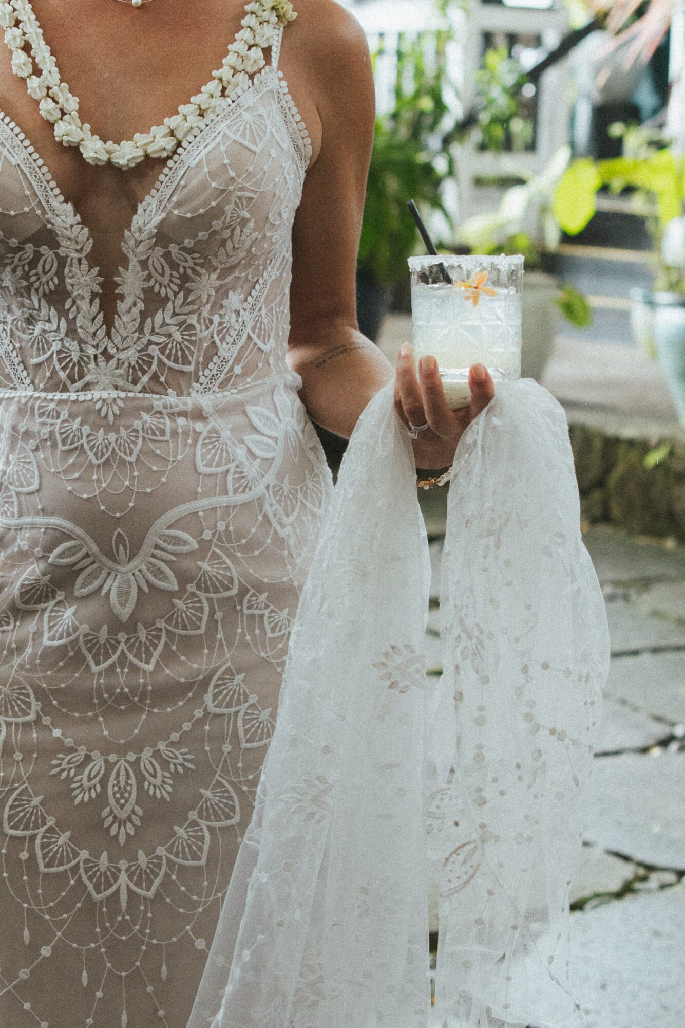 Person in a lace dress holding a glass with a drink, outdoors with greenery and a vehicle in the background.