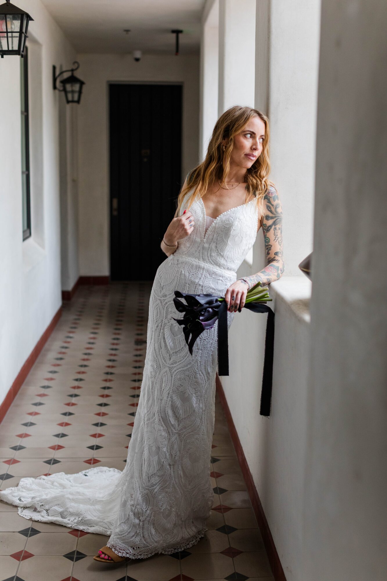 Woman in a white lace dress standing in a hallway, holding a bouquet, looking out a window.