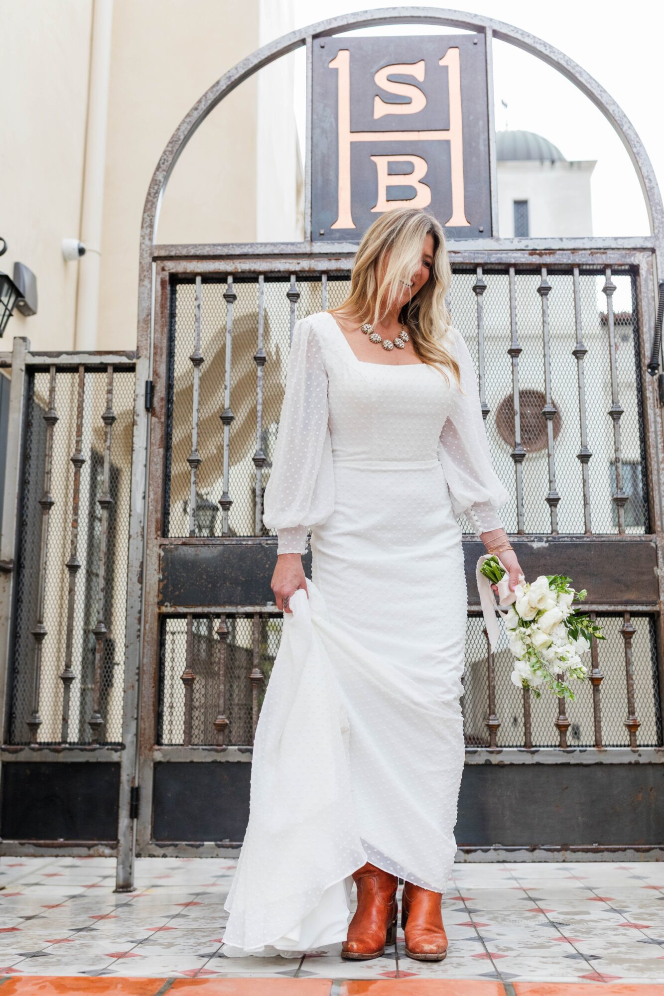 Woman in white dress holding bouquet, standing in front of a gate with a sign above, on a tiled sidewalk.