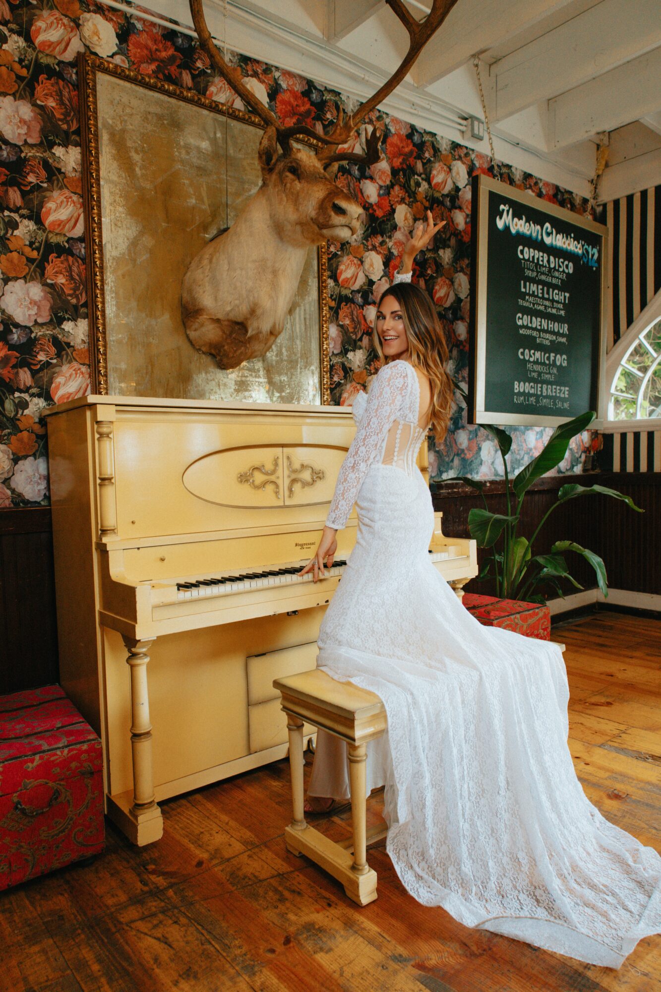 Woman in white dress sitting on a piano bench, with a mounted animal head and floral wallpaper behind her.