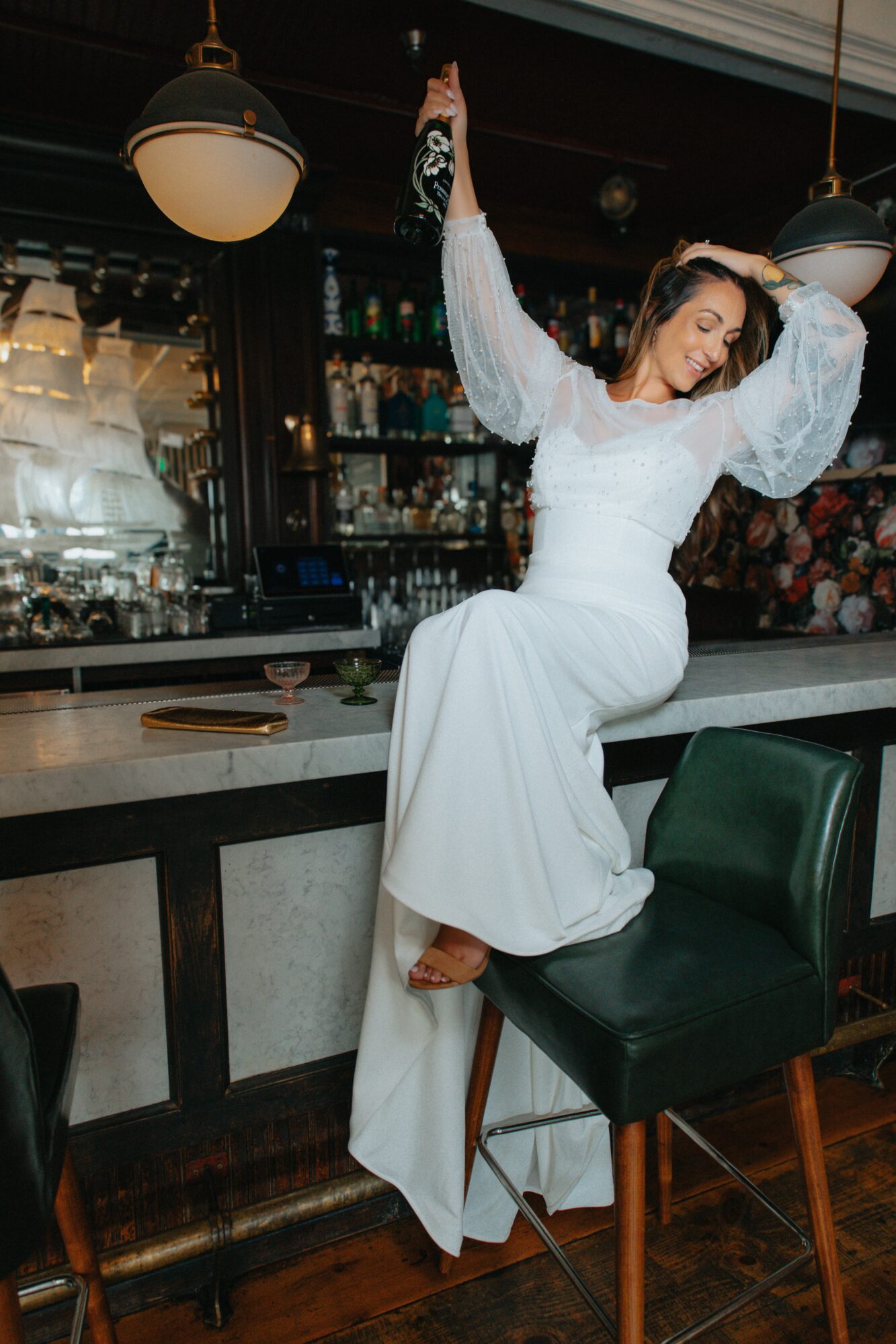 Woman in white dress sitting on bar stool at bar counter, holding a bottle, in a dimly lit bar with shelves of bottles behind her.