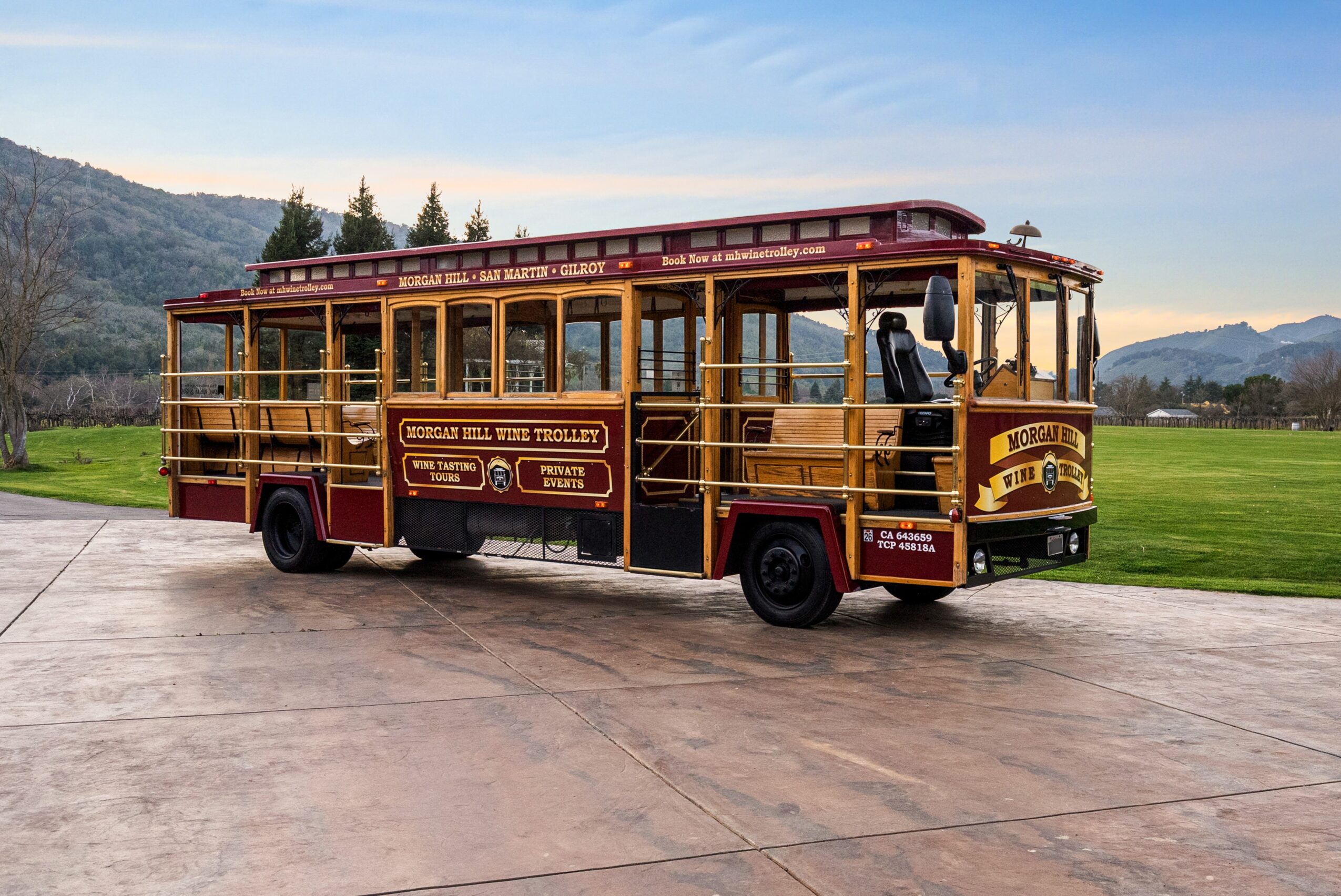 Vintage trolley bus with wooden accents parked on a paved area, with green fields and mountains in the background.