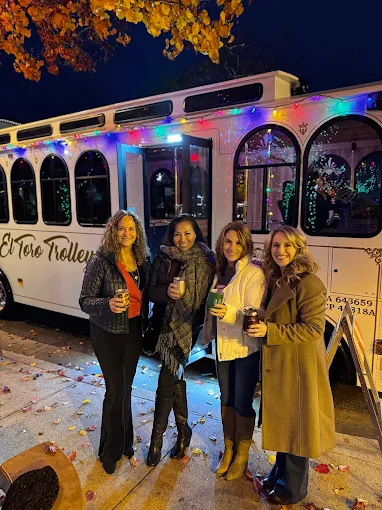 Four women stand in front of a decorated bus at night, holding drinks and smiling, with colorful lights and autumn leaves nearby.