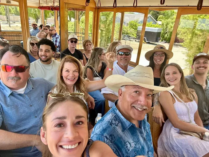 Group of people smiling inside a wooden structure with large windows, outdoors with trees visible outside, sunny day.