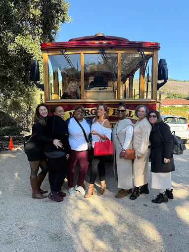 Group of six women standing in front of a red and yellow trolley bus outdoors, with trees and a building in the background.