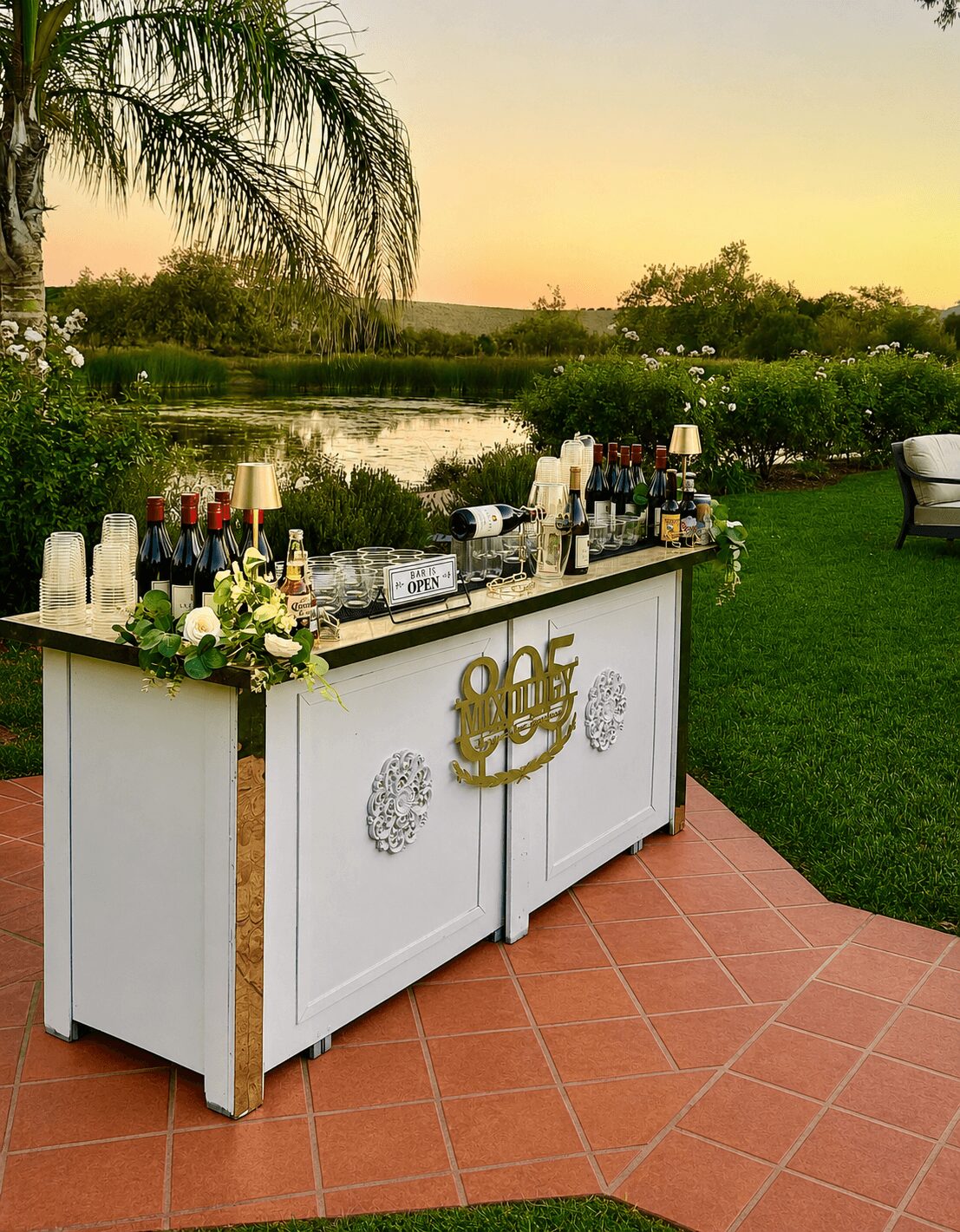 Outdoor bar setup with bottles, flowers, and a sign, on a tiled patio near a lake at sunset, with trees and grass.