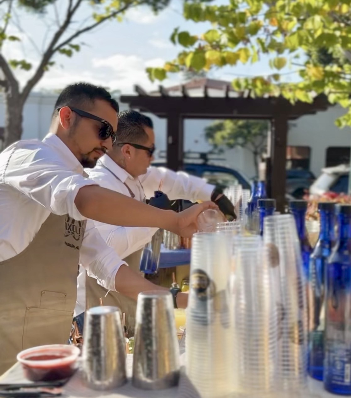 Two men in white shirts and aprons preparing drinks outdoors with cups and bottles, trees and a building in background.