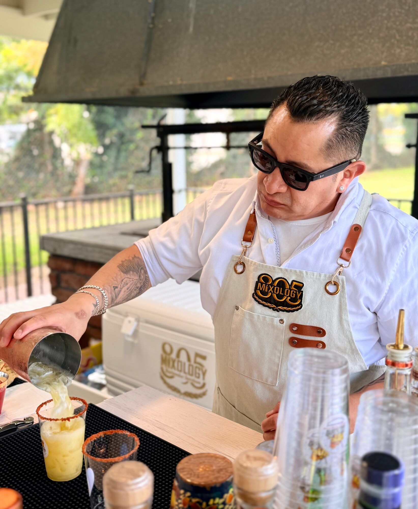 Person wearing sunglasses and apron preparing a drink outdoors, with a fence and trees in background.