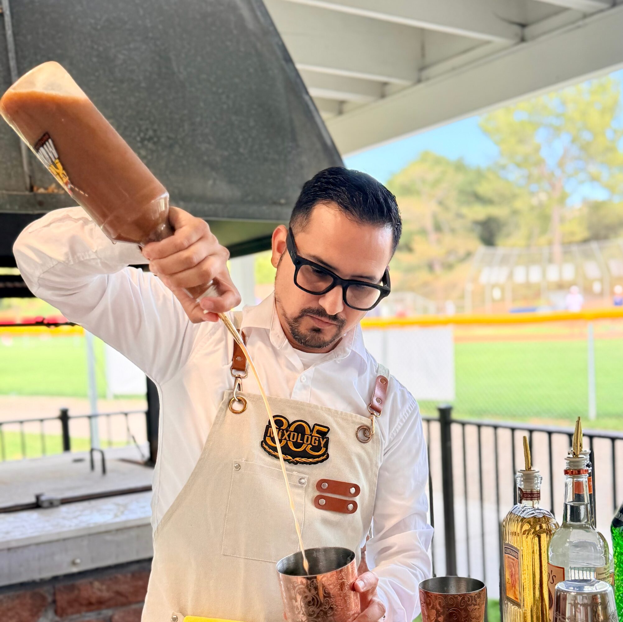 Bartender pouring a drink into a copper mug outdoors, with bottles on the bar and a grassy field in the background.
