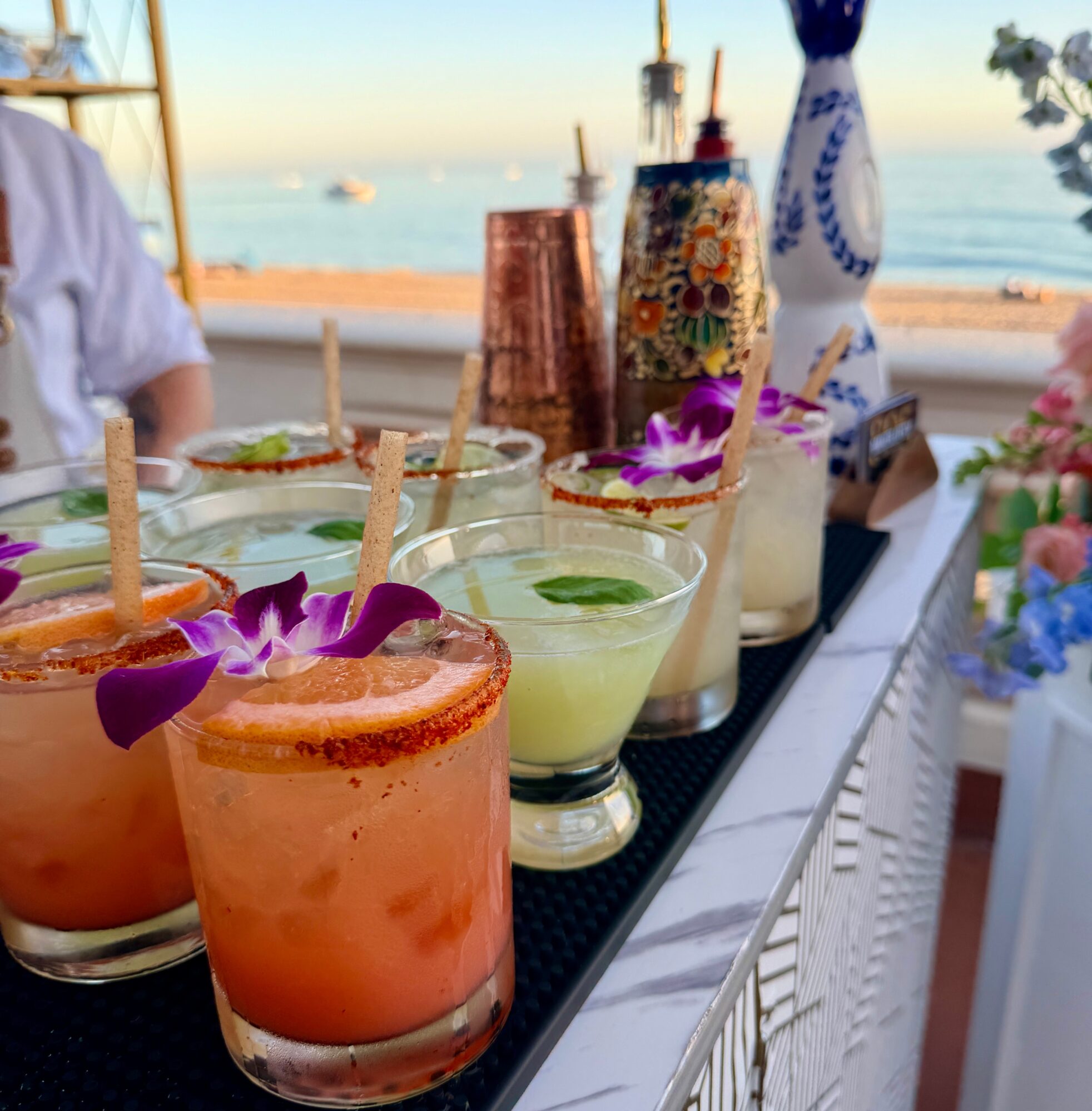 Colorful cocktails with garnishes on a table near the beach, with boats and water in the background.