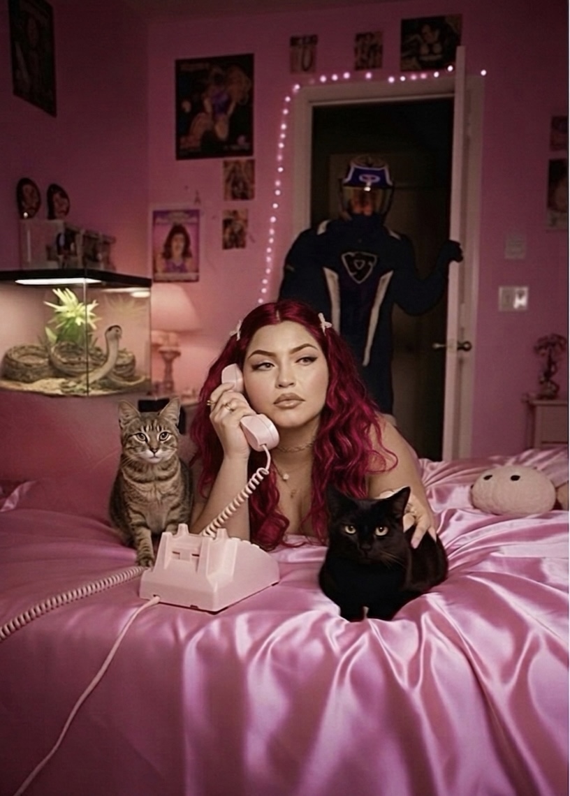 Young woman with red hair and makeup, sitting on pink bed with two cats, in a decorated pink bedroom.