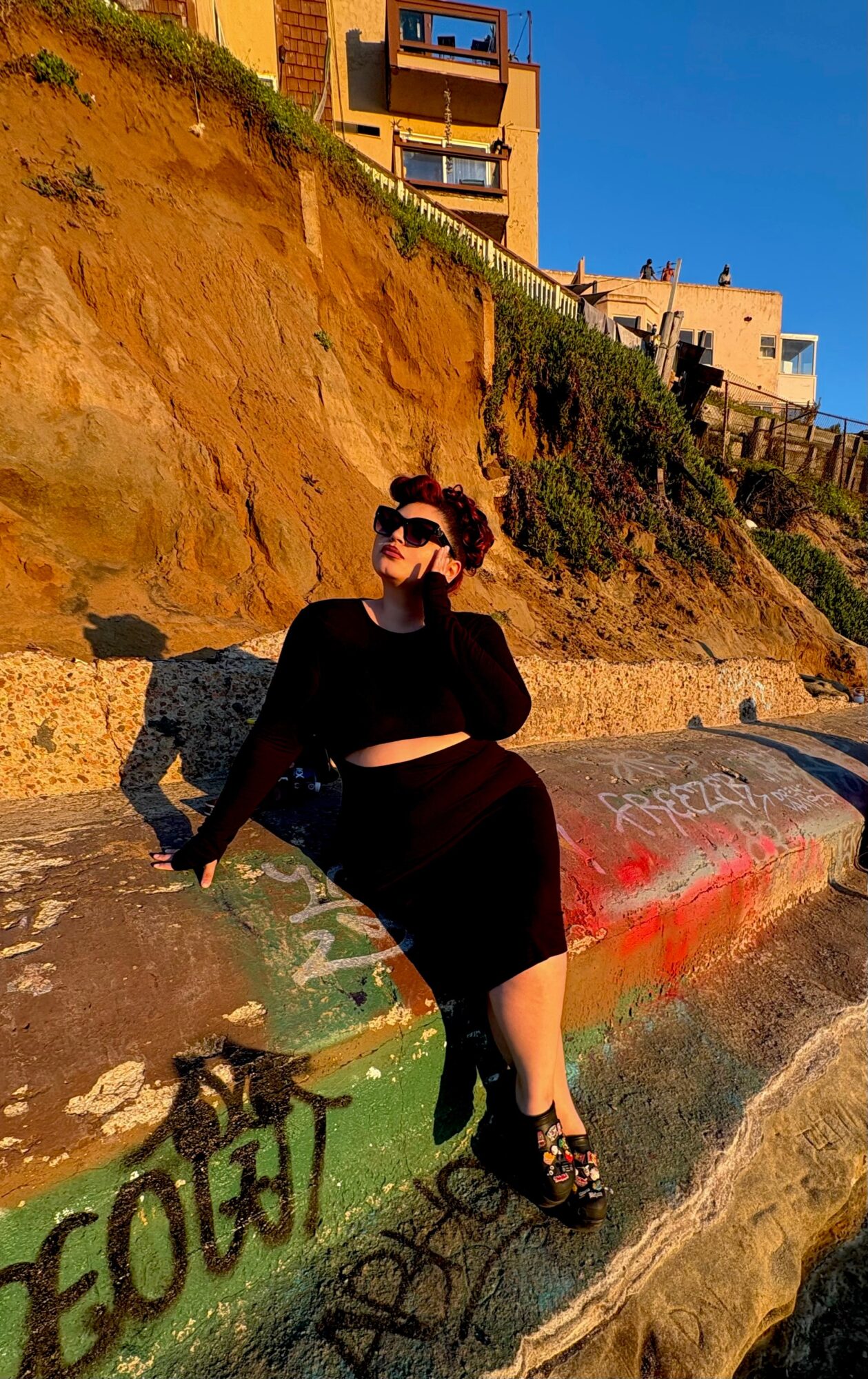 Woman in black dress and sunglasses sitting on a graffiti-covered concrete surface outdoors, with a hillside and buildings in the background.