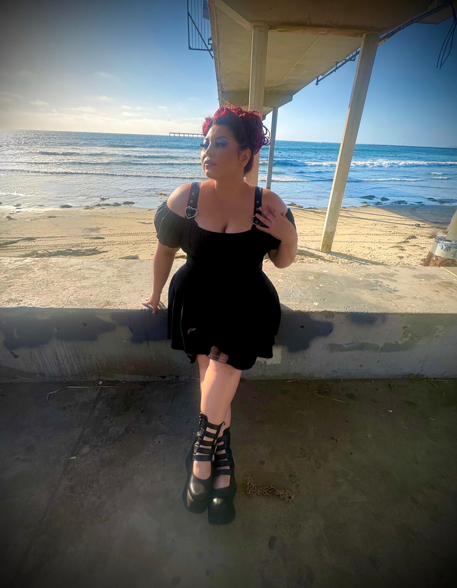 Woman in black dress with platform shoes standing on beach under a structure, ocean and sky in background.